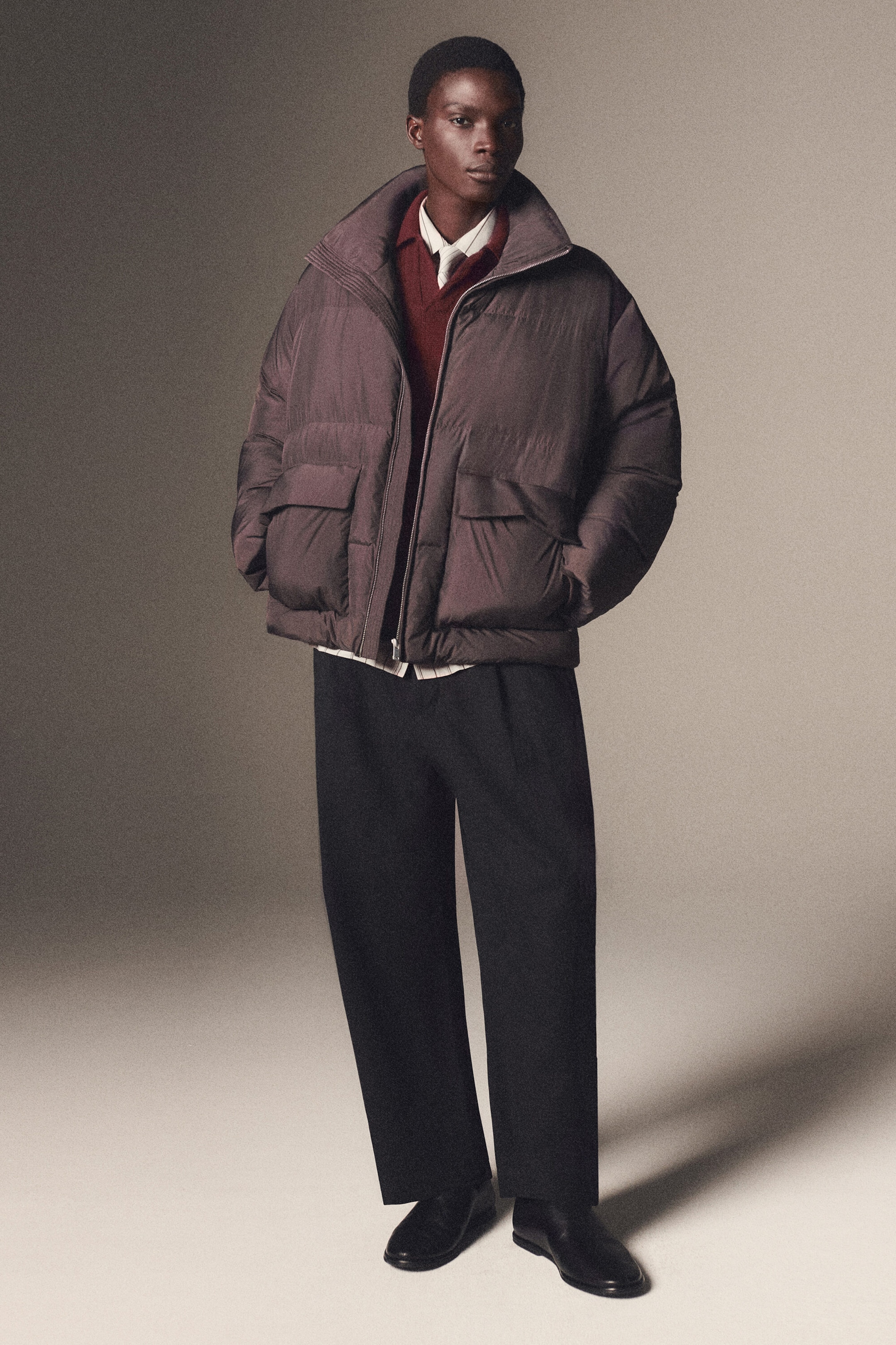The funnel-neck nylon down puffer jacket is shown from the front in a relaxed, oversized fit in a dark taupe hue. It features horizontal quilting, a high funnel collar, and large front flap pockets, and is worn open over a red knit vest and a collared shirt to highlight its layered styling. The funnel-neck nylon down puffer jacket is shown from the front in a relaxed, oversized fit in a dark taupe hue. It features horizontal quilting, a high funnel collar, and large front flap pockets, and is worn open over a red knit vest and a collared shirt to highlight its layered styling.