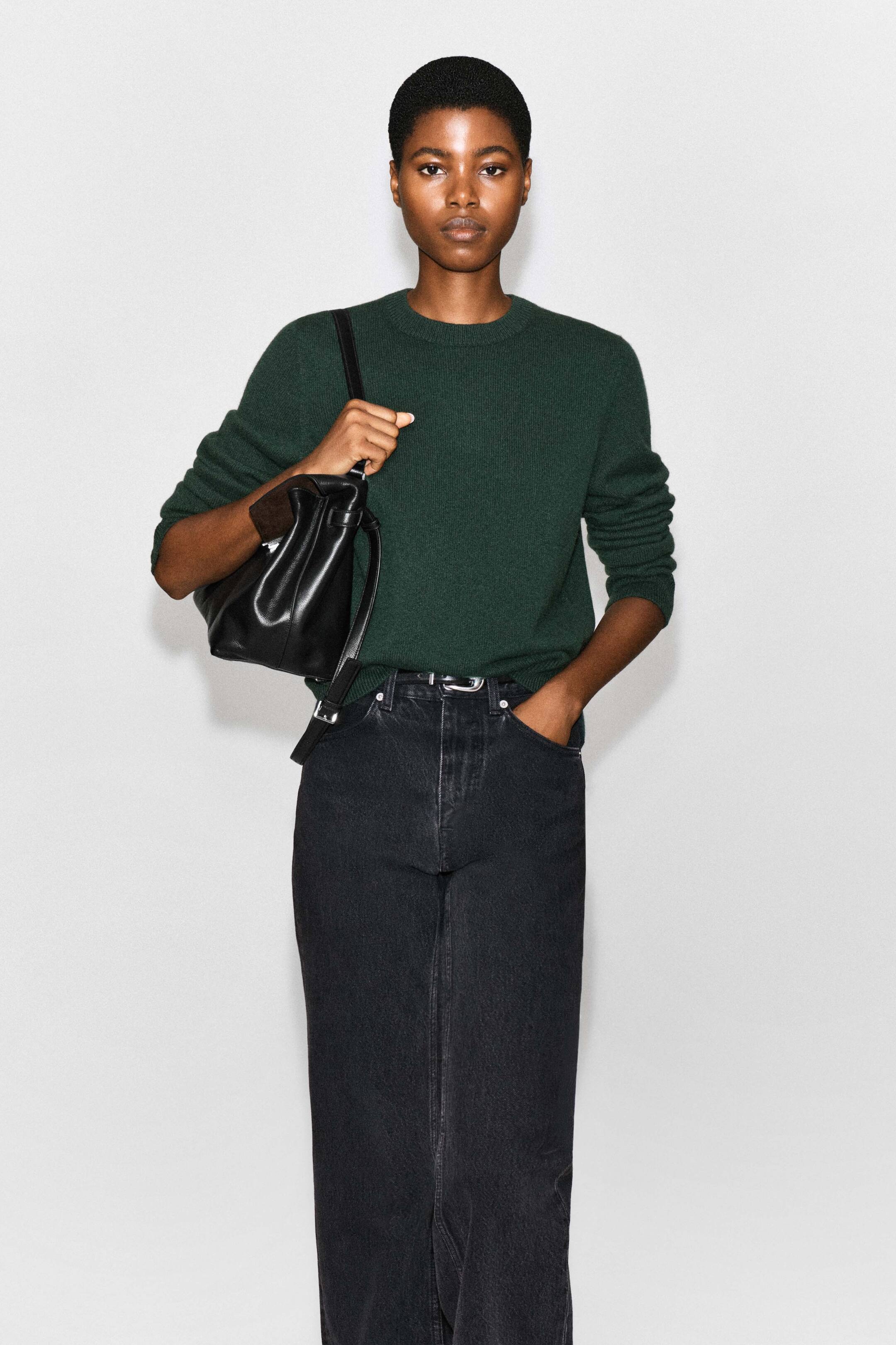 View larger image: A dark green cashmere crew-neck jumper is being worn by the model, tucked into high-waisted black denim jeans. The jumper features ribbed trims at the collar and cuffs, with its sleeves slightly pushed up, and is styled with a black leather shoulder bag.