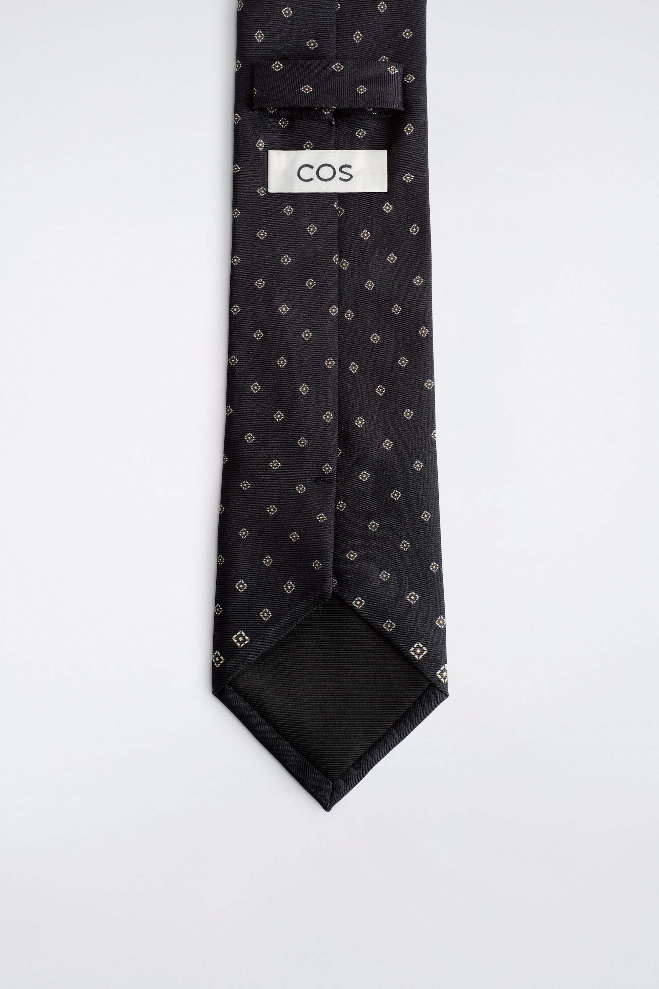 A dark-coloured tie is shown from the back, displaying a small geometric foulard pattern, a self-fabric keeper loop, and a white brand label printed with "COS". The pointed end reveals a black ribbed lining. A dark-coloured tie is shown from the back, displaying a small geometric foulard pattern, a self-fabric keeper loop, and a white brand label printed with "COS". The pointed end reveals a black ribbed lining.