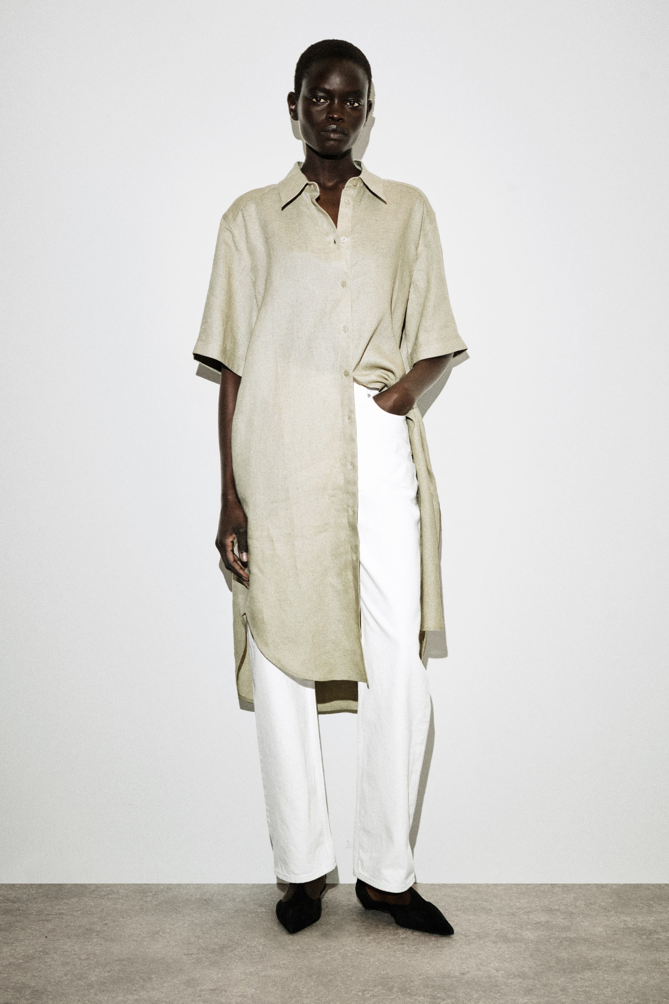 Größeres Bild ansehen: Ein langes, kastiges Leinenhemd in hellem Beige wird vom Modell getragen. Dieses locker geschnittene Kleidungsstück verfügt über einen spitzen Kragen, kurze Ärmel und einen Knopfverschluss vorne, der über die Hüften hinaus mit einem abgerundeten Saum und Seitenschlitzen reicht. Das Modell steht und hat eine Hand in der Tasche der weißen Hose, die unter dem Hemd getragen wird.