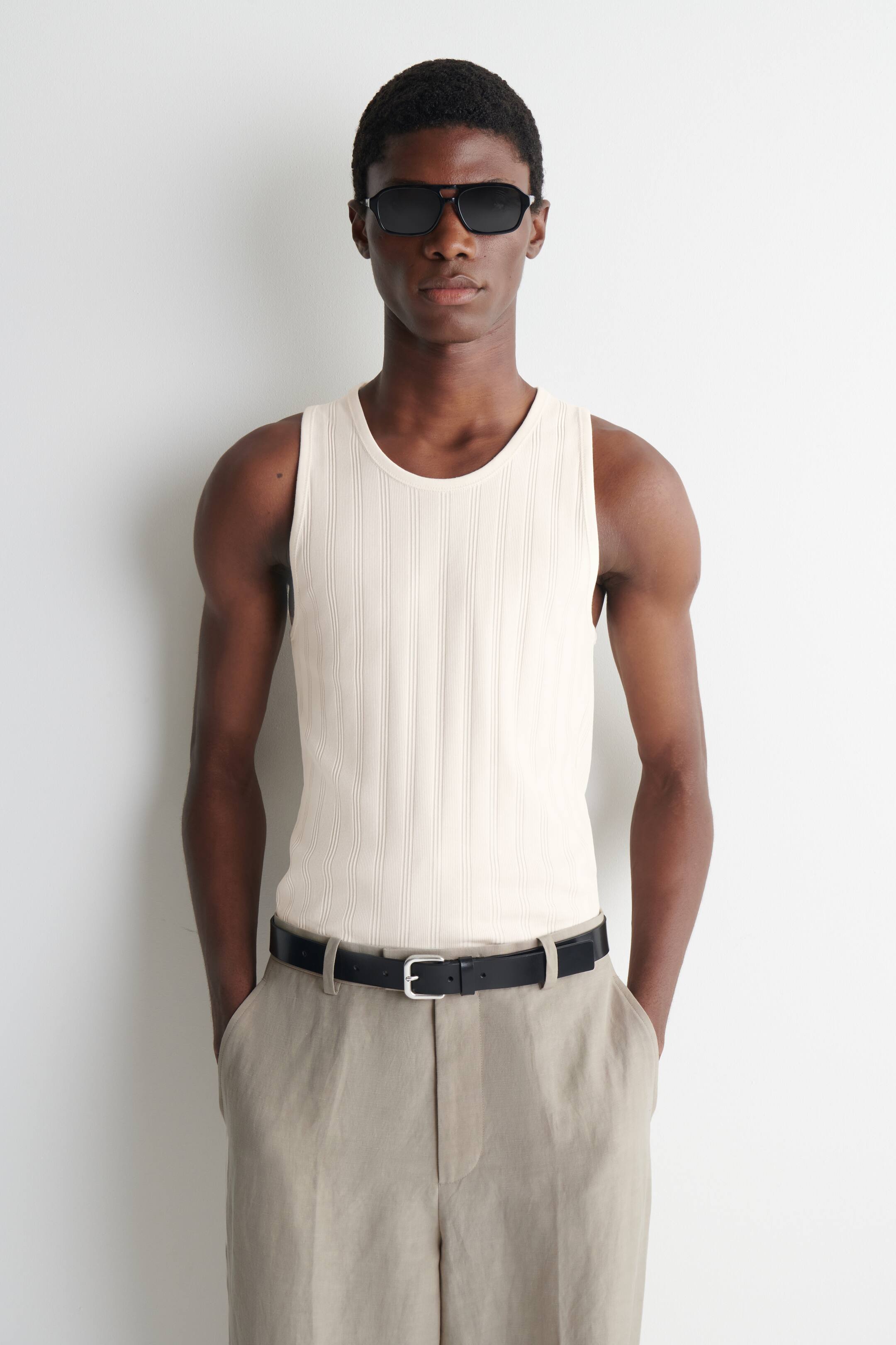 Visa större bild: En krämfärgad tanktopp med breda ribbor och rund hals samt en strömlinjeformad, smal passform bärs av modellen. Tanktoppen är stylad med ljusa byxor och ett svart bälte.