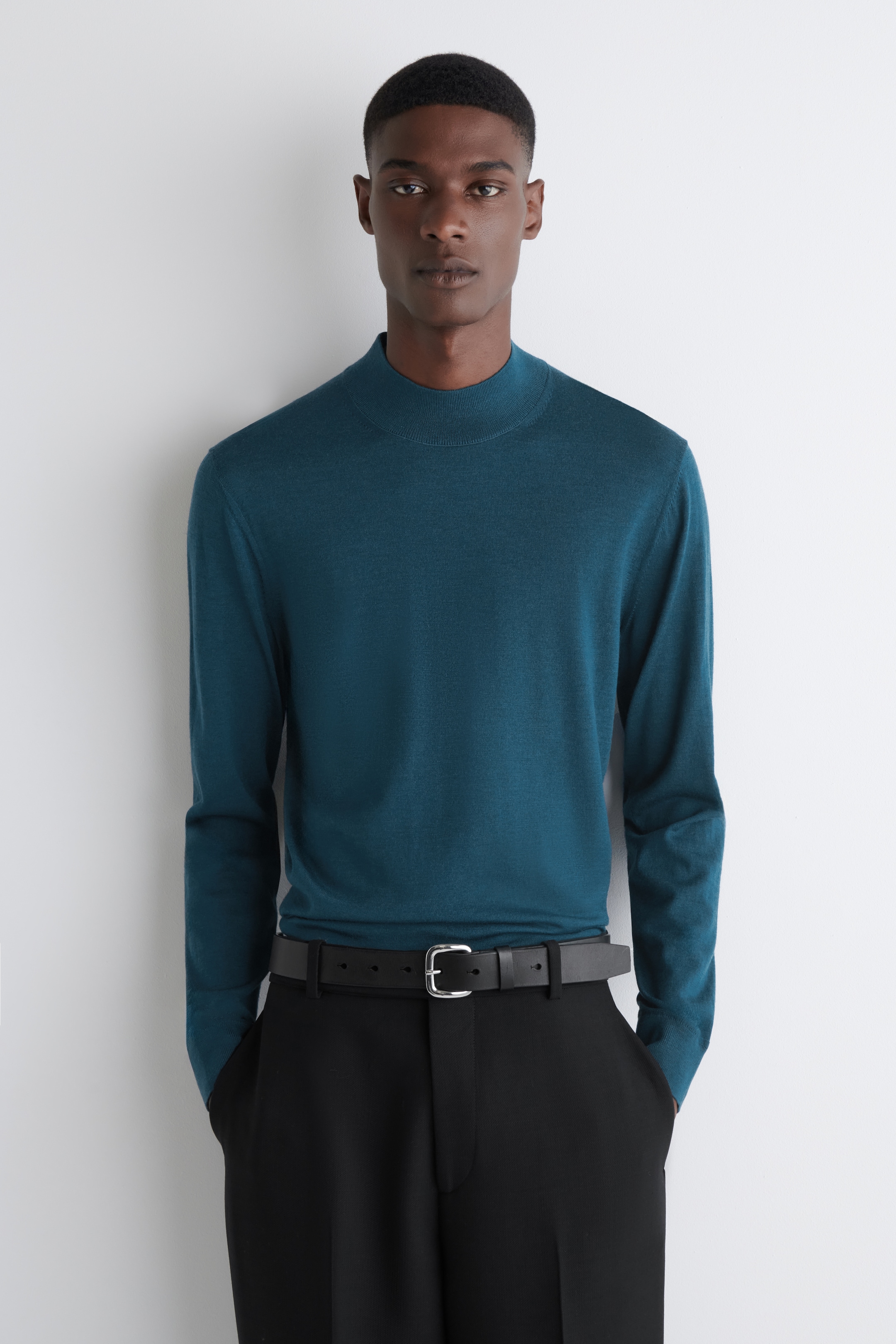 View larger image: A teal mock neck sweater is being presented from a front-facing view, showcasing its smooth, fine knit and long sleeves. It is featuring a regular fit and is being worn tucked into black trousers.