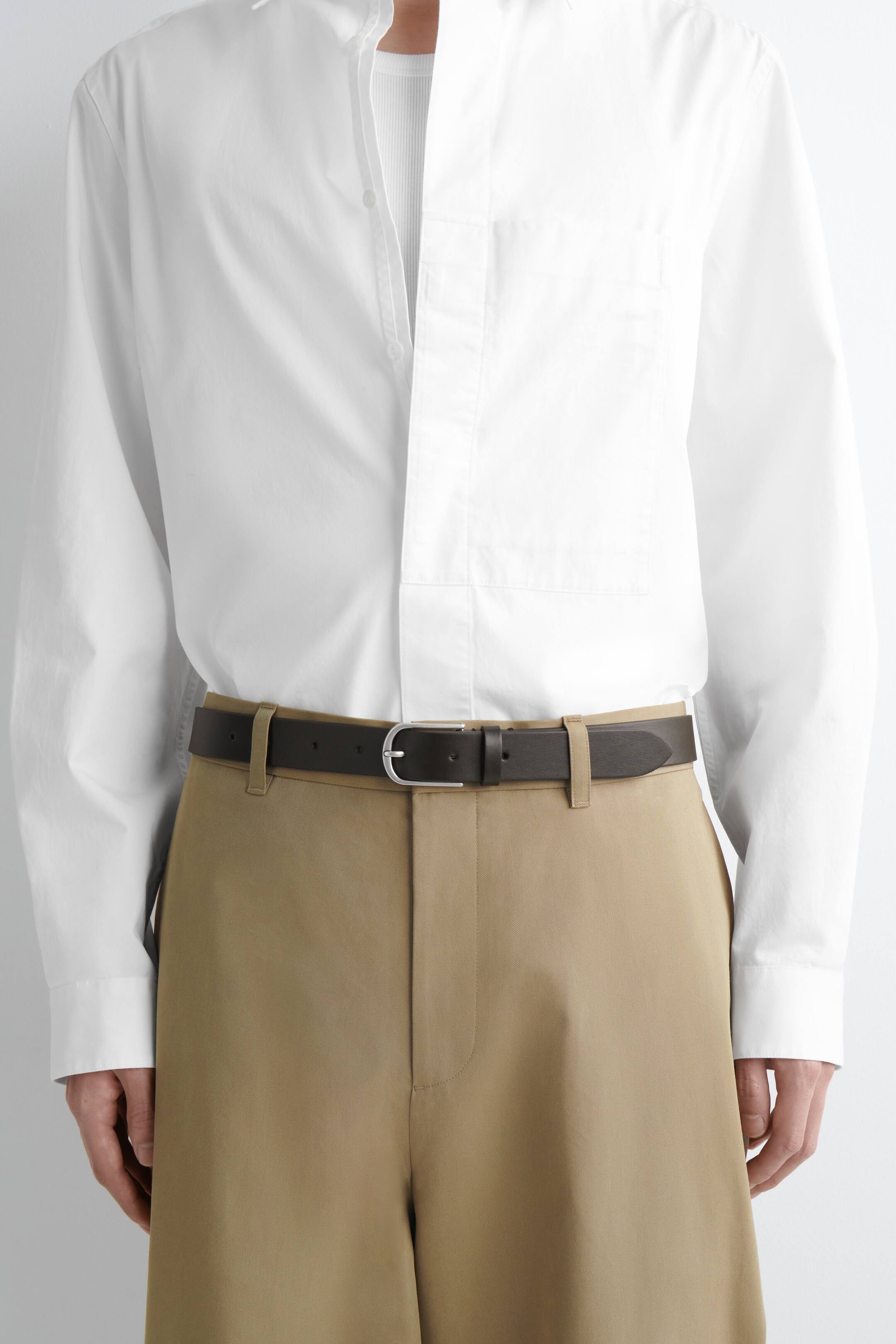 Преглед на по-голямо изображение: Тъмнокафяв кожен колан се носи около талията, закопчан със сребристо, заоблено катарама. Коланът е представен отпред, като преминава през гайките на каки панталони и е насложен върху бяла риза.