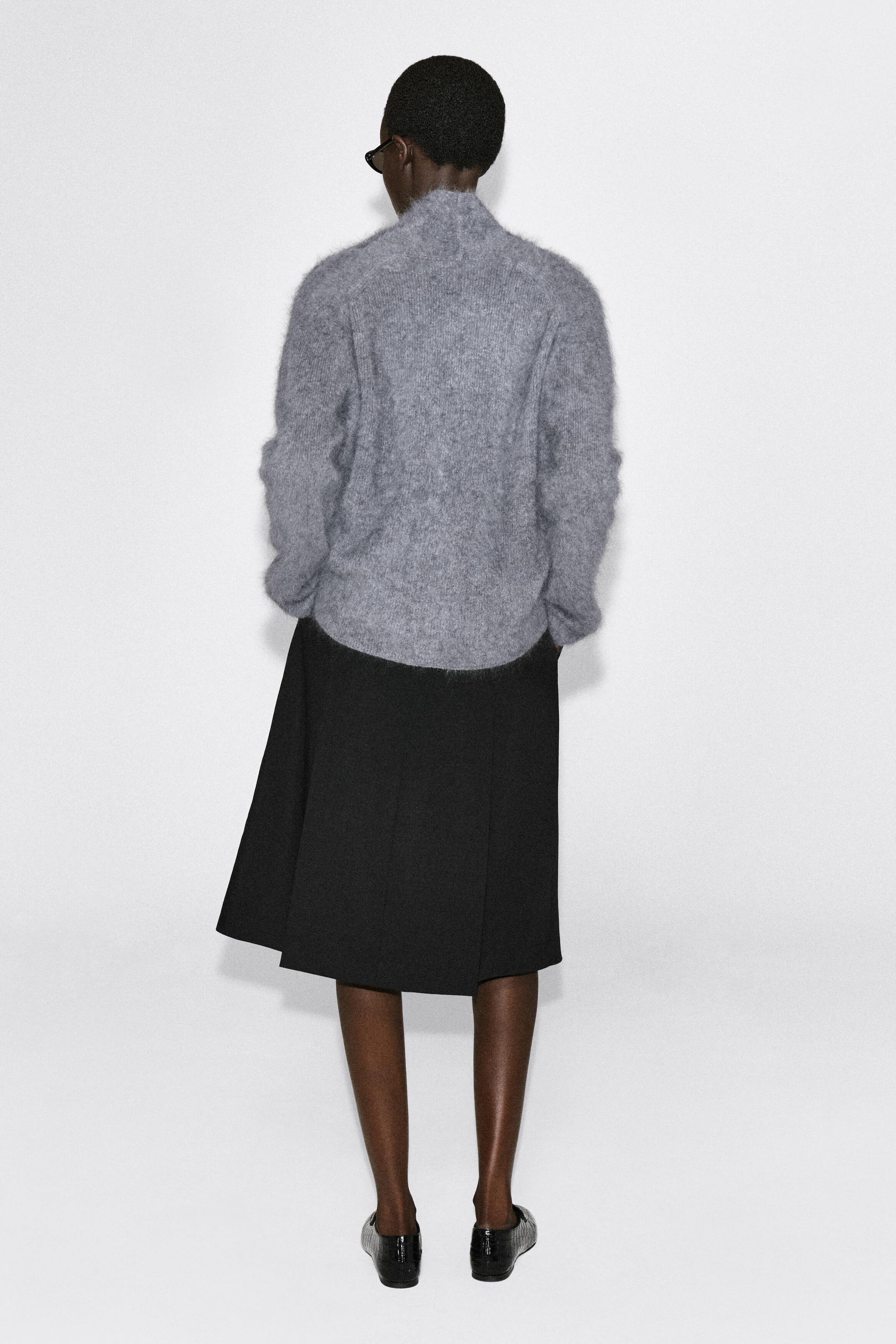 The draped brushed merino wool cardigan is shown from the back, highlighting its soft, fuzzy texture and relaxed silhouette. It features raglan sleeves, a high collar, and a ribbed hem resting over a black skirt.