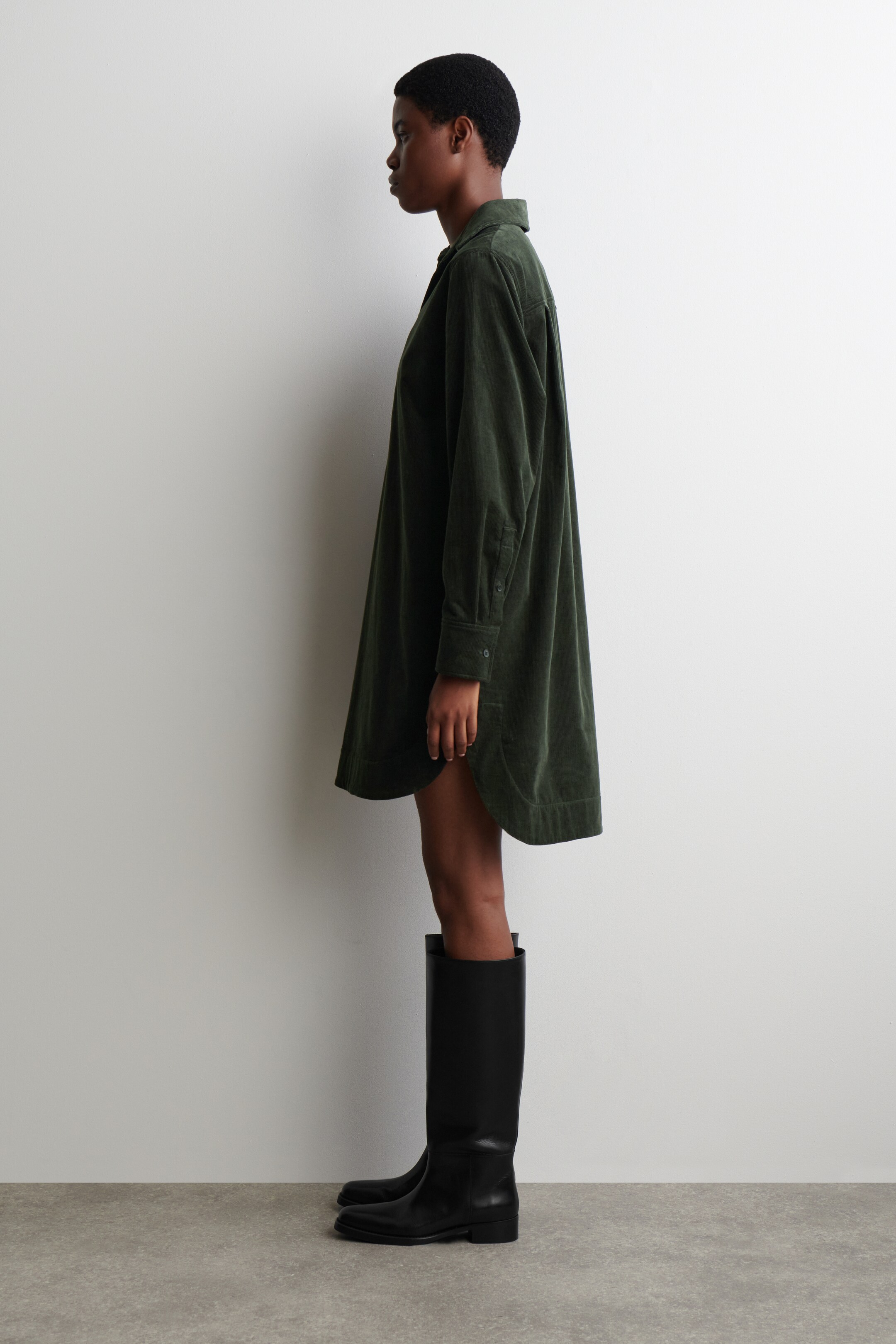 Vezi imaginea mai mare: O rochie-cămașă din catifea raiată verde închis este vizibilă dintr-o vedere laterală, având un guler clasic, mâneci lungi cu manșete cu nasturi și un tiv rotunjit, mai lung în spate, care cade deasupra genunchilor și este asortată cu cizme negre până la genunchi.