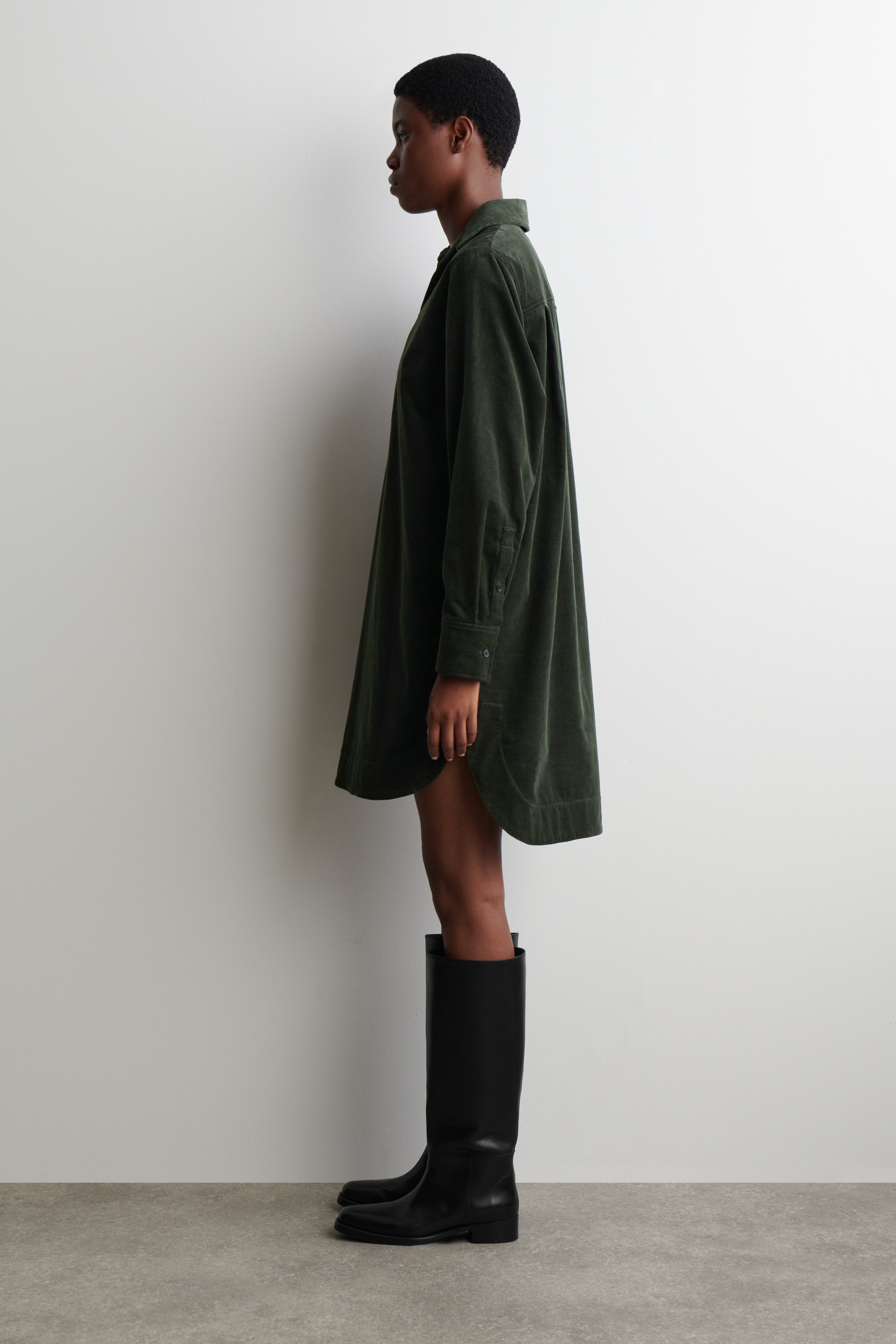 Visualizza immagine più grande: Un abito a camicia in velluto a coste verde scuro è visibile di profilo, con un colletto classico, maniche lunghe con polsini abbottonati, e un orlo arrotondato e alto-basso che cade sopra le ginocchia e viene abbinato a stivali neri alti fino al ginocchio.