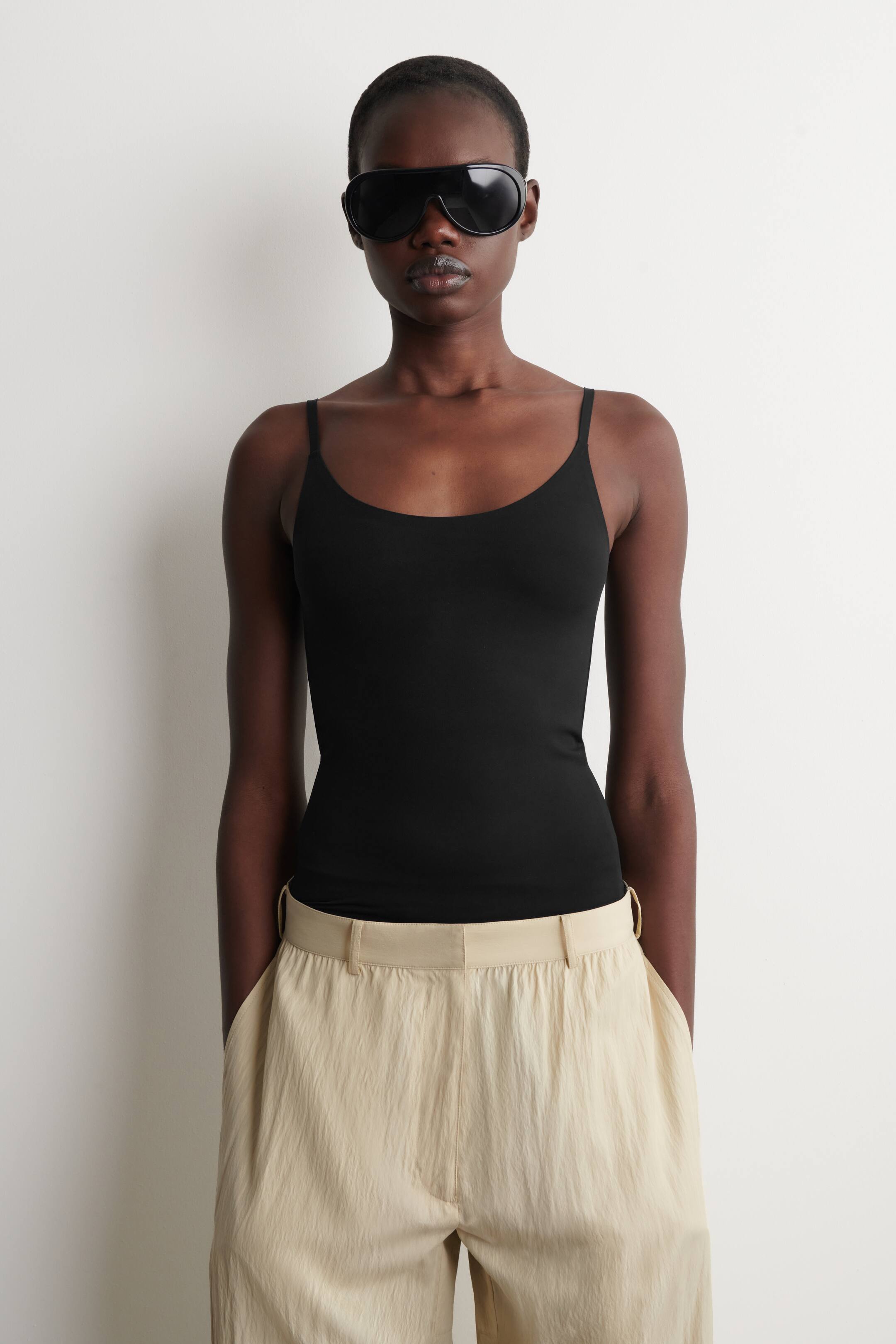 Ver imagem maior: Uma camisola preta com alças finas em estilo espaguete e um decote redondo ajusta-se suavemente ao torso. A bainha está inserida em calças de cor clara, exibindo todo o seu comprimento.