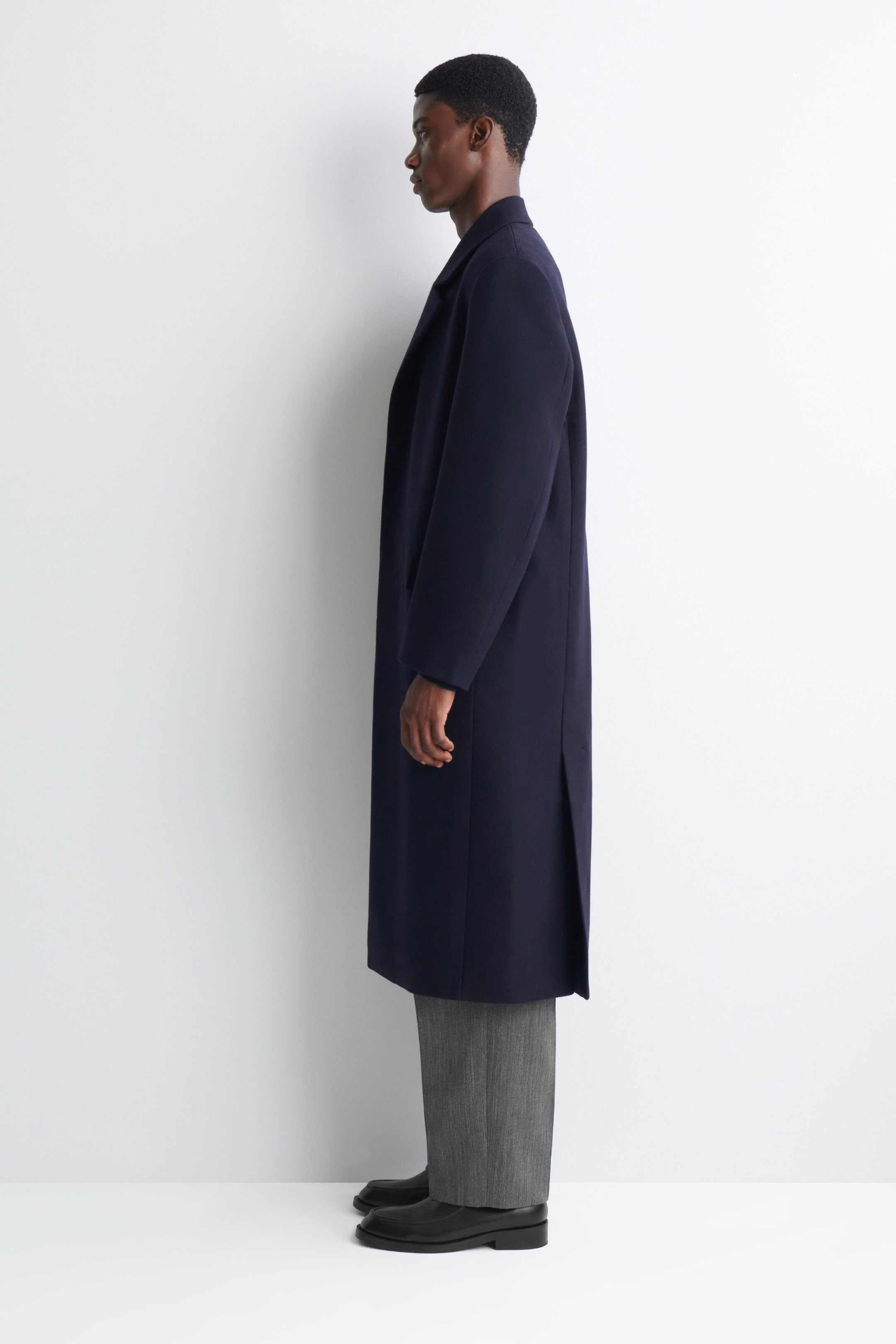 The model is wearing a navy long coat with notched lapels and a back vent, layered over grey trousers. The model is wearing a navy long coat with notched lapels and a back vent, layered over grey trousers.