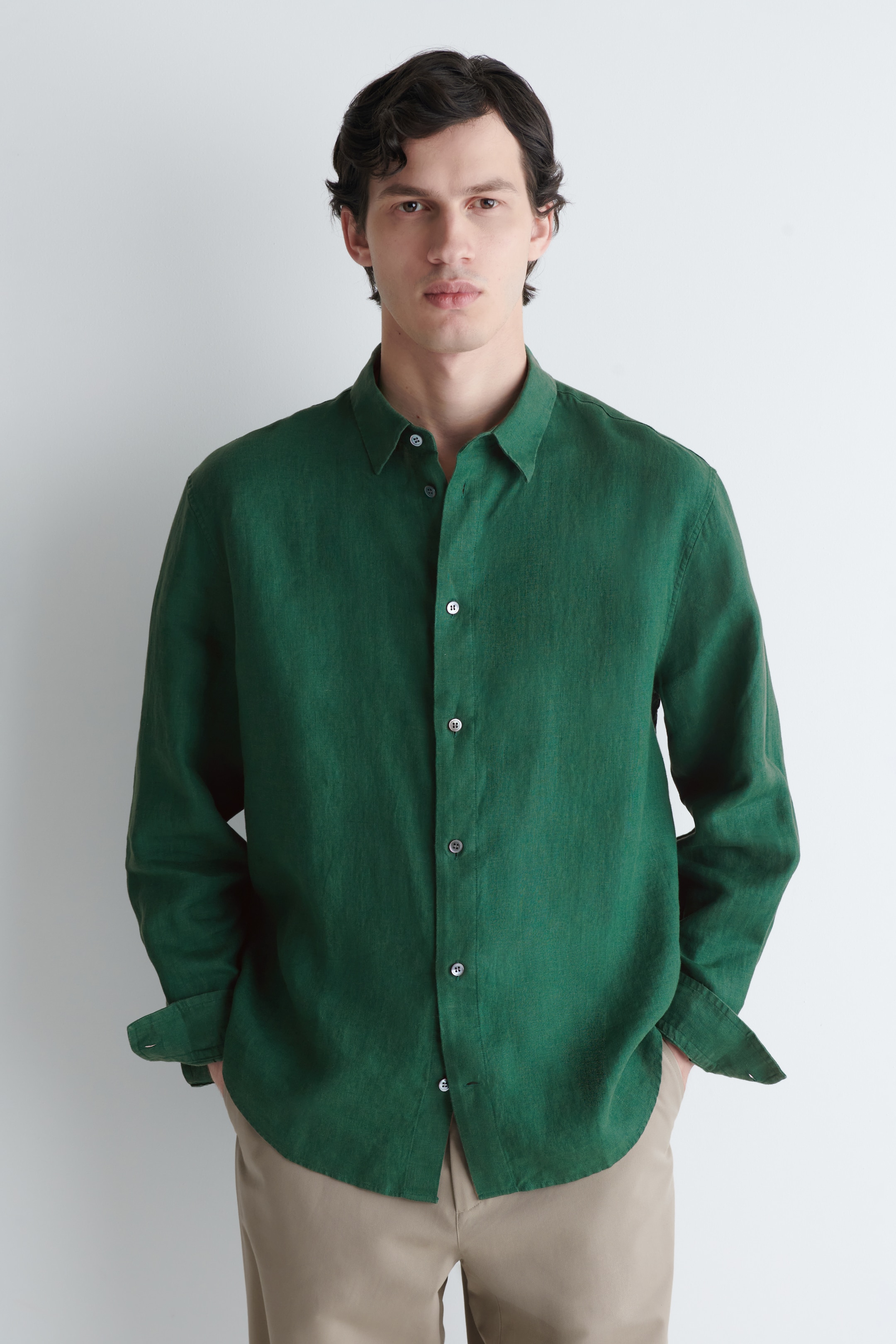 הצגת תמונה גדולה יותר: חולצה ירוקה כהה עם מרקם, בעלת כפתורים בחזית, מוצגת כשהיא פונה קדימה, עם צווארון מחודד, כפתורים בצבע בהיר לאורך החזית ושרוולים ארוכים עם חפתים, כאשר אחד מהם מעט מקופל. גם שולי החולצה המעוגלים נראים לעין.