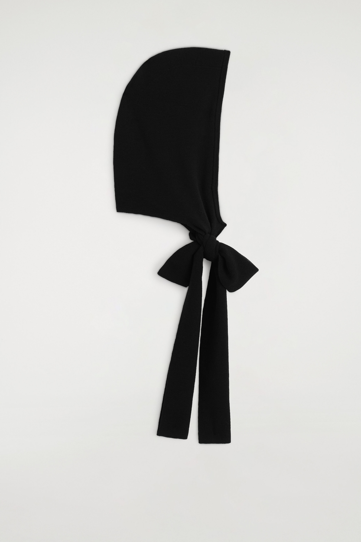 A black knit hooded scarf is shown in right-side profile, highlighting its structured hood shape and fine-knit texture. The long straps are tied into a bow at the base of the hood, with two flat ends hanging straight down.