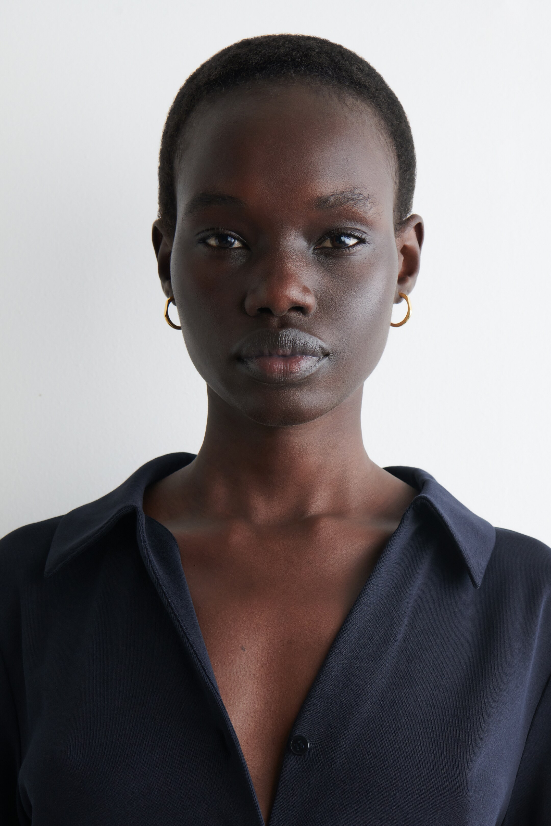 Agrandir l'image: Une chemise bleu marine foncé à col est exposée, avec un décolleté en V ouvert et un bouton visible. Deux petites boucles d'oreilles en anneau de ton doré sont également portées.