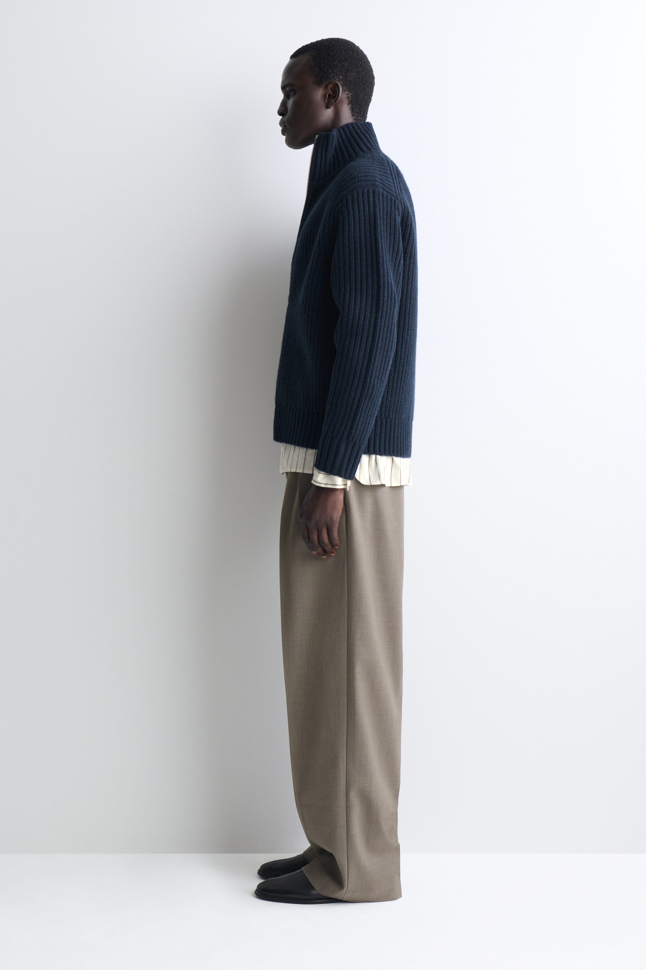 A navy blue ribbed-knit zip-up cardigan is worn facing left, featuring a high collar, ribbed cuffs, and a straight hem. It is layered over a light-colored striped shirt and paired with wide-leg taupe trousers that drape over black leather shoes. A navy blue ribbed-knit zip-up cardigan is worn facing left, featuring a high collar, ribbed cuffs, and a straight hem. It is layered over a light-colored striped shirt and paired with wide-leg taupe trousers that drape over black leather shoes.