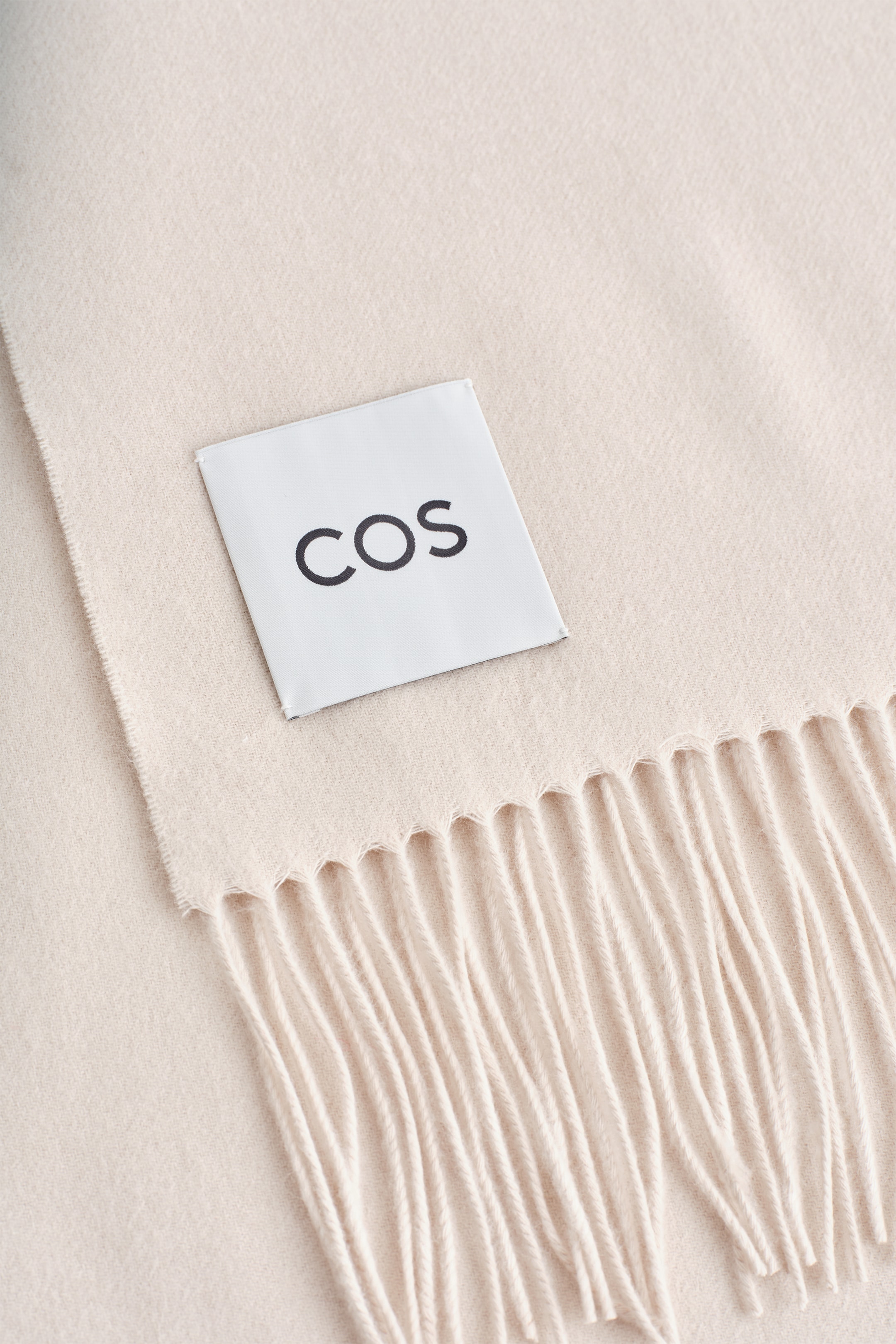 Agrandir l'image: Une écharpe de couleur crème est présentée en gros plan. Une étiquette rectangulaire blanche montre le logo 'COS' cousu sur la surface de l'écharpe, et des bords à franges apparaissent à une extrémité.