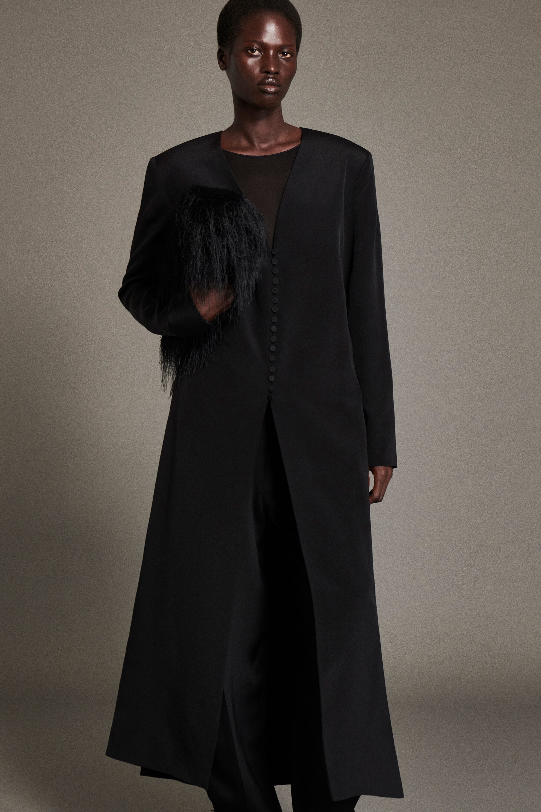 A model facing forward is wearing a black button-detail silk-crepe long coat featuring a collarless V-neckline, structured shoulders, and a row of small covered buttons that opens into a high front slit. The coat is styled over matching black pleated silk-crepe barrel-leg pants and a black top, with a textured black faux-fur accessory held under the arm.