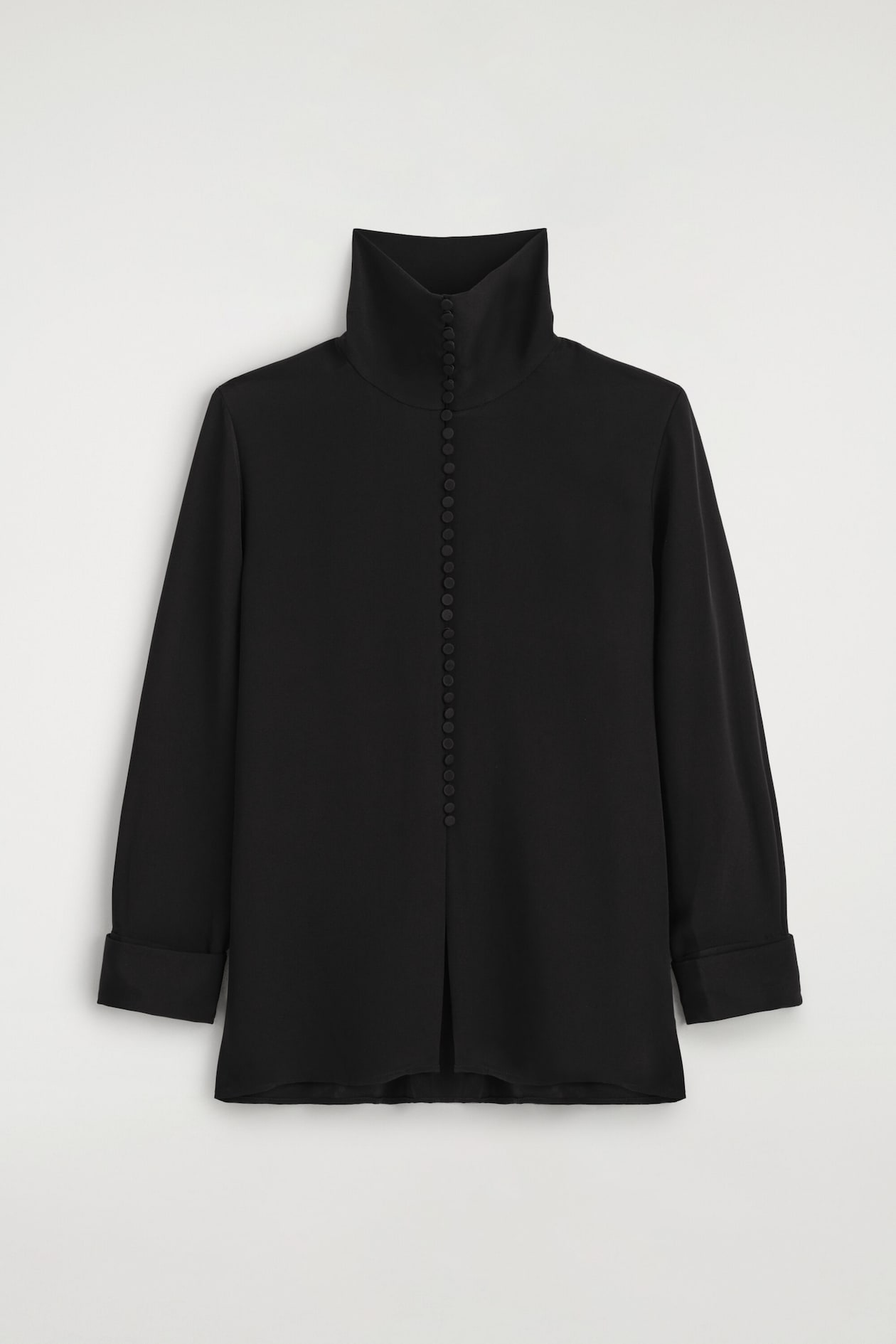 A front-facing black silk shirt showcases a high, structured funnel neck and a vertical row of small, fabric-covered buttons running down the centre. The shirt features long sleeves with wide folded cuffs and a relaxed silhouette with a small central slit at the hem.