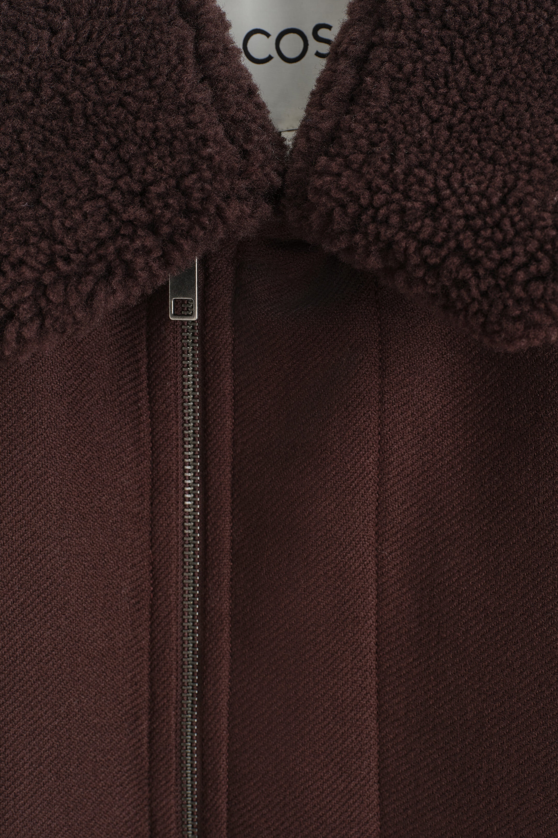 A dark brown jacket is shown in a close-up front view, featuring a plush, textured shearling collar and a diagonal twill-weave fabric body. A silver-tone metal zipper with a rectangular pull tab runs down the front, with a white brand label partially visible at the inner collar. A dark brown jacket is shown in a close-up front view, featuring a plush, textured shearling collar and a diagonal twill-weave fabric body. A silver-tone metal zipper with a rectangular pull tab runs down the front, with a white brand label partially visible at the inner collar.
