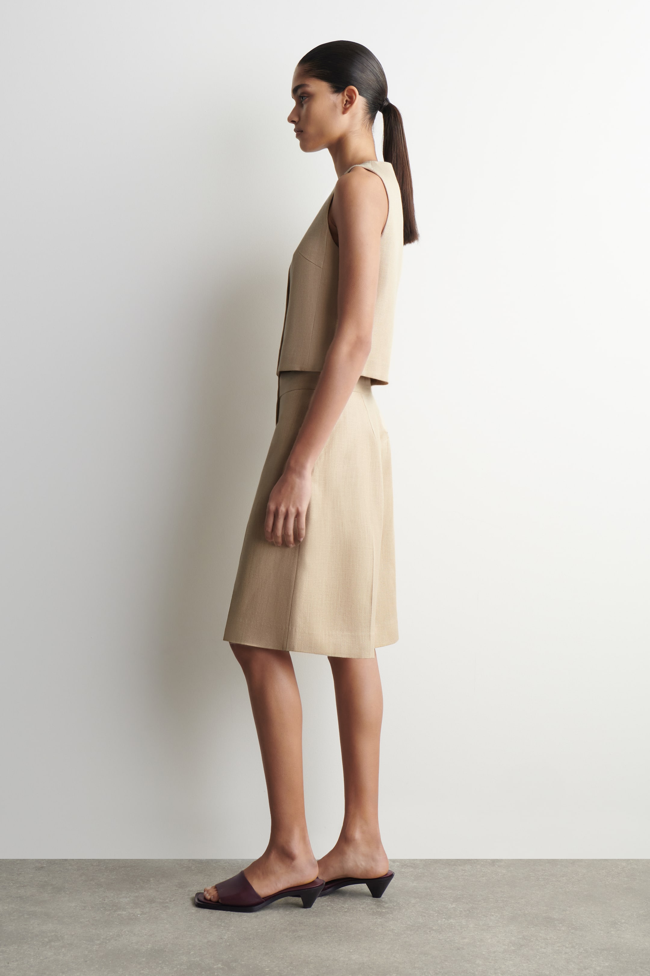 הצגת תמונה גדולה יותר: אפוד קצר ללא שרוולים בצבע חום בהיר וחצאית תואמת באורך ברך בגזרה A מוצגים בפרופיל צדדי, כאשר האפוד כולל סגירת כפתורים בחזית והחצאית יושבת במותן הטבעית. כפכפים פתוחים בצבע חום כהה עם עקבים נמוכים ומתרחבים משלימים את המראה המתואם.