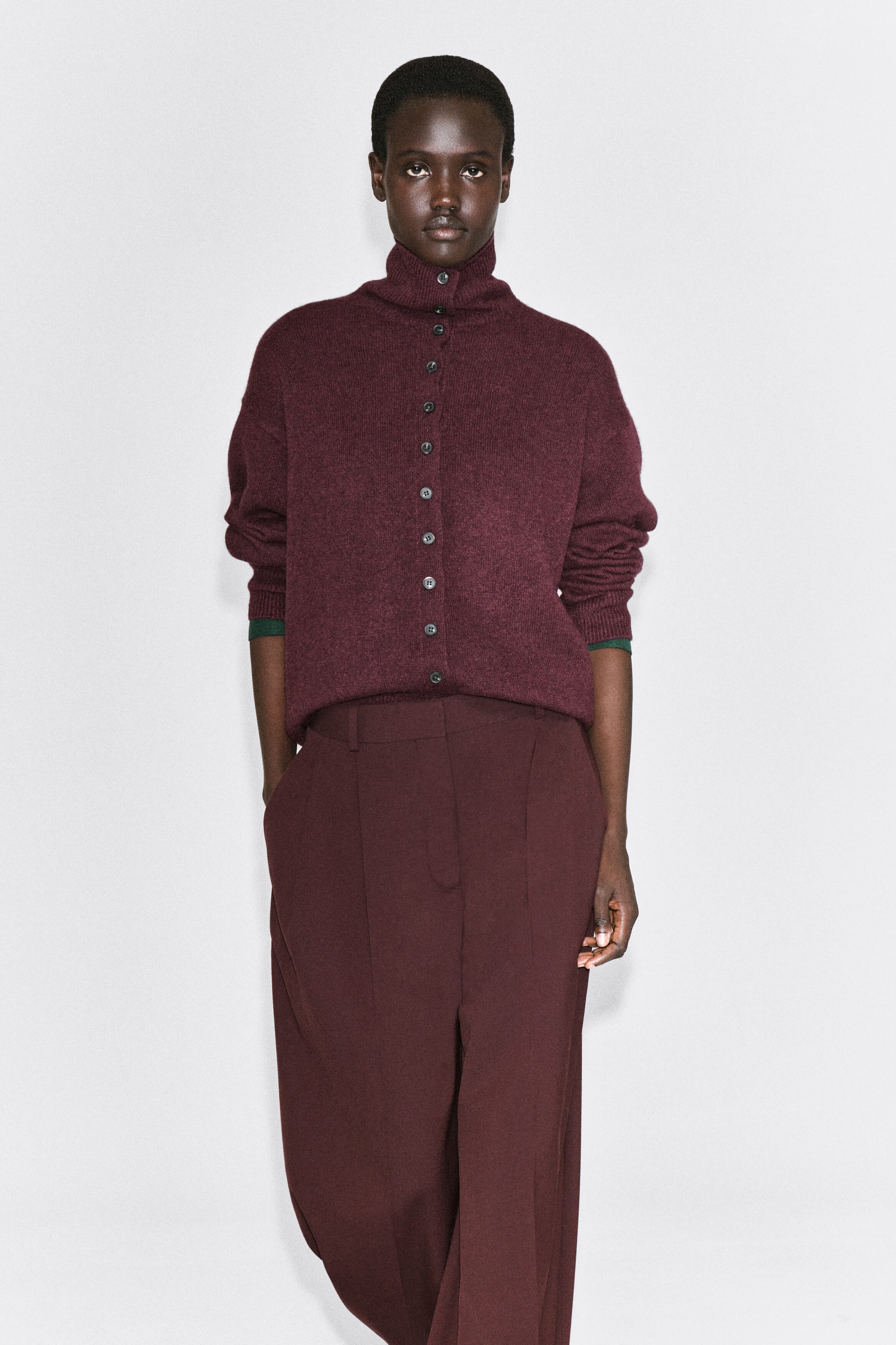 A model is wearing a burgundy cashmere cardigan, facing front, featuring a button-up front that extends into a high funnel neckline. The relaxed-fit cardigan has dropped shoulders with ribbed cuffs and is styled tucked into matching wide-leg trousers. A model is wearing a burgundy cashmere cardigan, facing front, featuring a button-up front that extends into a high funnel neckline. The relaxed-fit cardigan has dropped shoulders with ribbed cuffs and is styled tucked into matching wide-leg trousers.