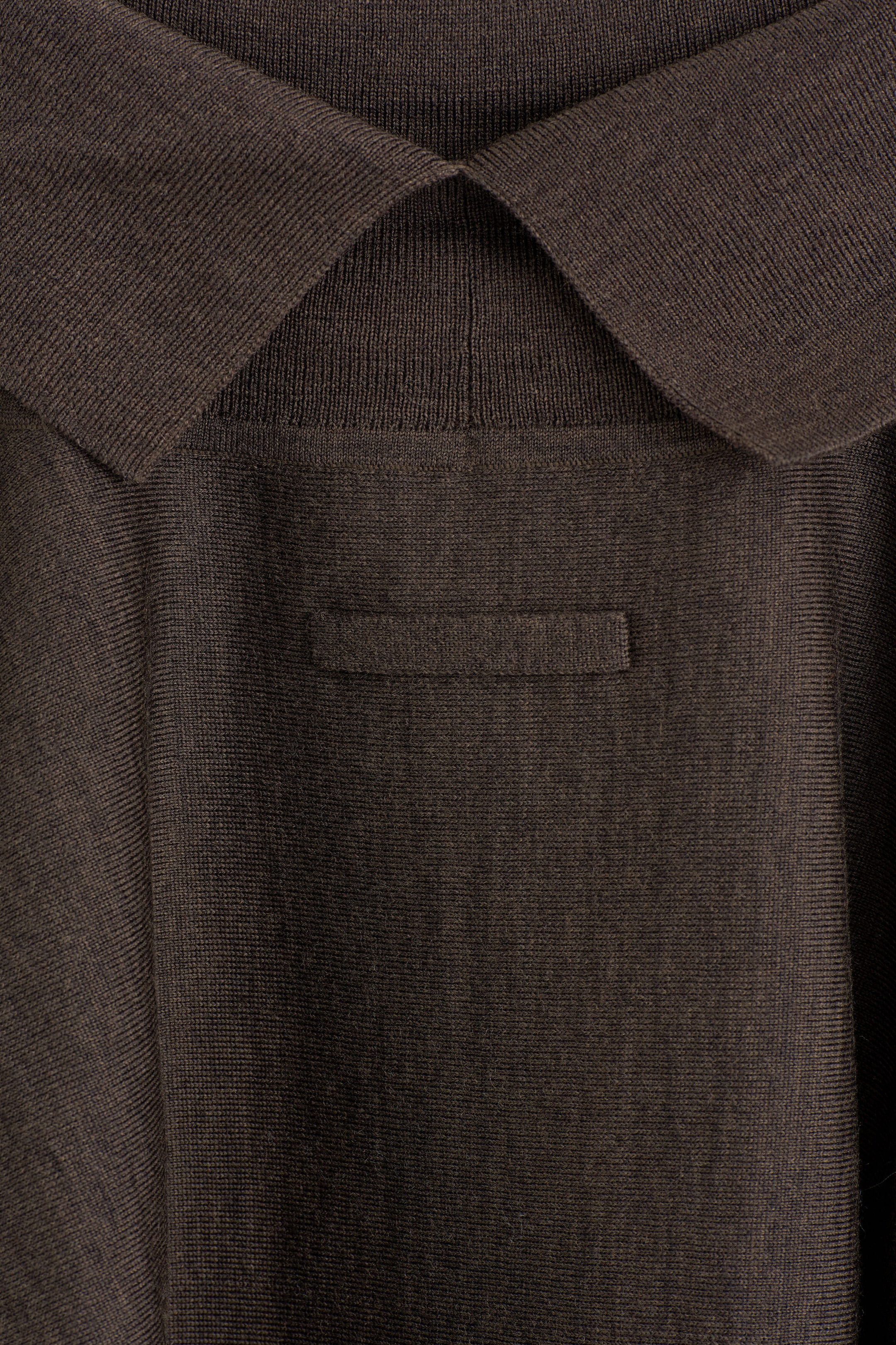 This dark brown knitted garment features a classic pointed collar and a small horizontal knitted tab detail below the back neckline. The fine-knit fabric displays a subtle, even texture. This dark brown knitted garment features a classic pointed collar and a small horizontal knitted tab detail below the back neckline. The fine-knit fabric displays a subtle, even texture.