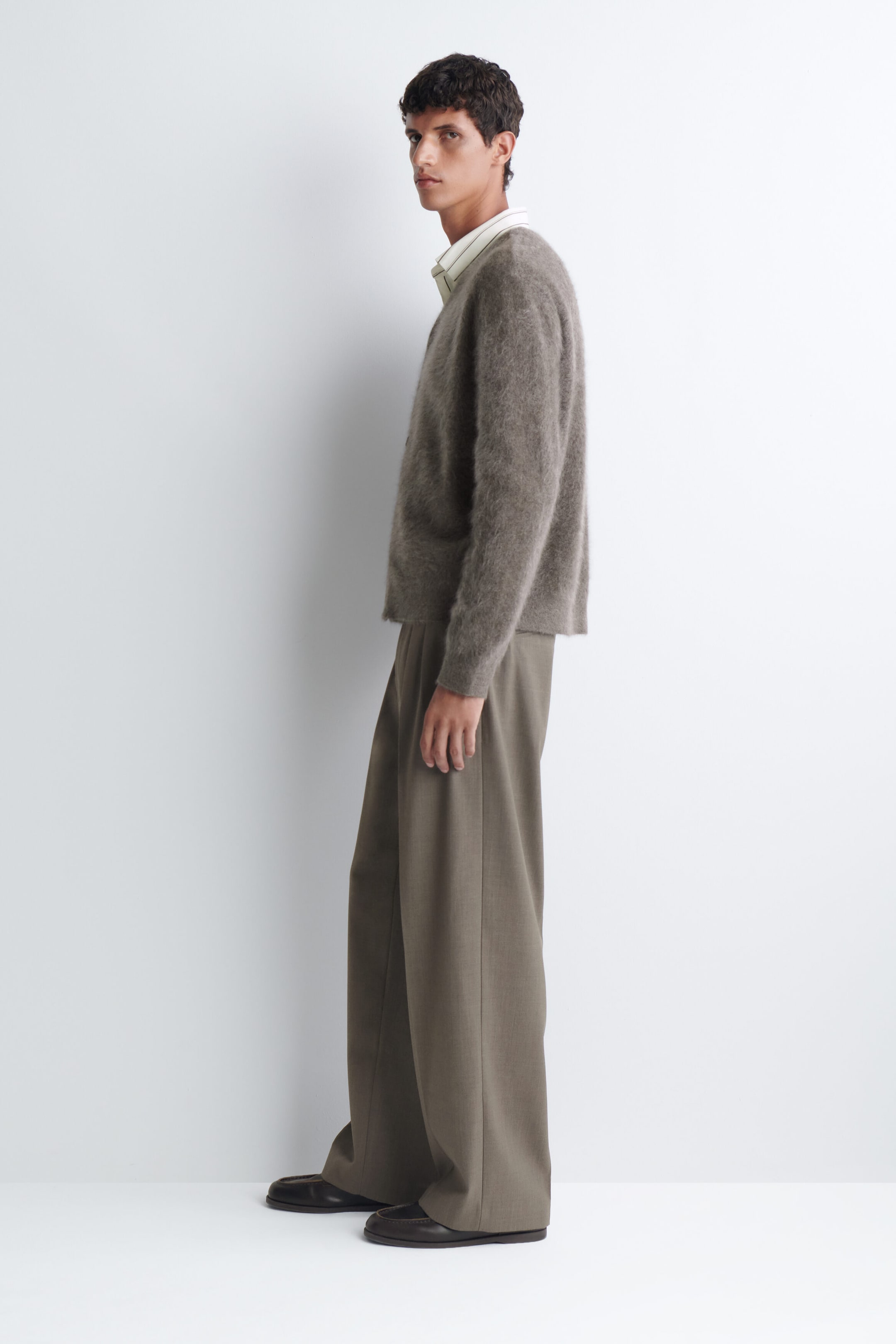 Un cardigan à col en V en cachemire brossé, dans une teinte taupe texturée, est porté par le mannequin par-dessus une chemise à rayures claires avec col. Le cardigan duveteux à manches longues est agencé avec un pantalon à jambe large et des mocassins foncés. Un cardigan à col en V en cachemire brossé, dans une teinte taupe texturée, est porté par le mannequin par-dessus une chemise à rayures claires avec col. Le cardigan duveteux à manches longues est agencé avec un pantalon à jambe large et des mocassins foncés.