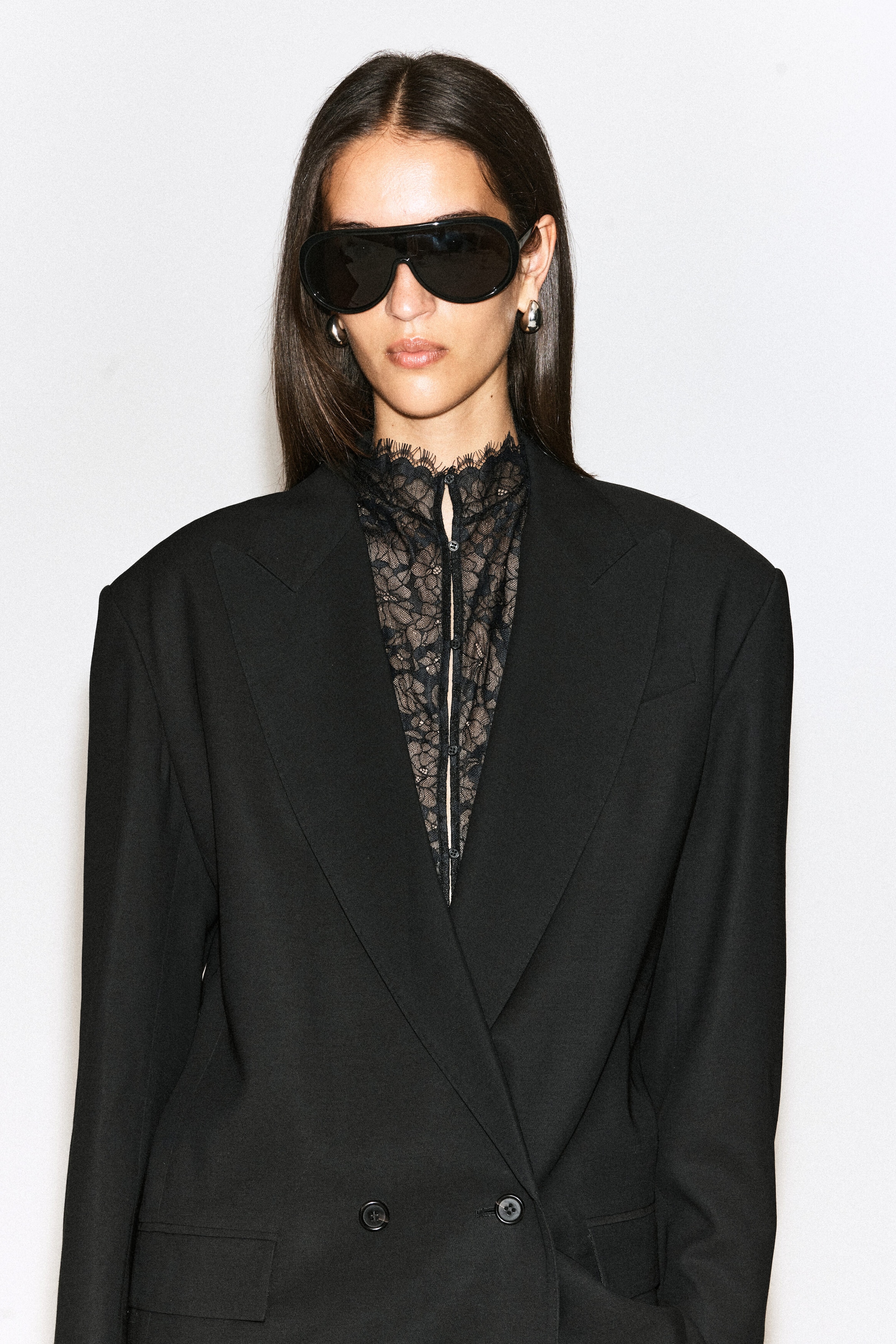 A black scalloped lace bib featuring a high neck with eyelash trim and a centre button closure is being worn by the model. The bib is layered underneath a structured, black double-breasted blazer. A black scalloped lace bib featuring a high neck with eyelash trim and a centre button closure is being worn by the model. The bib is layered underneath a structured, black double-breasted blazer.