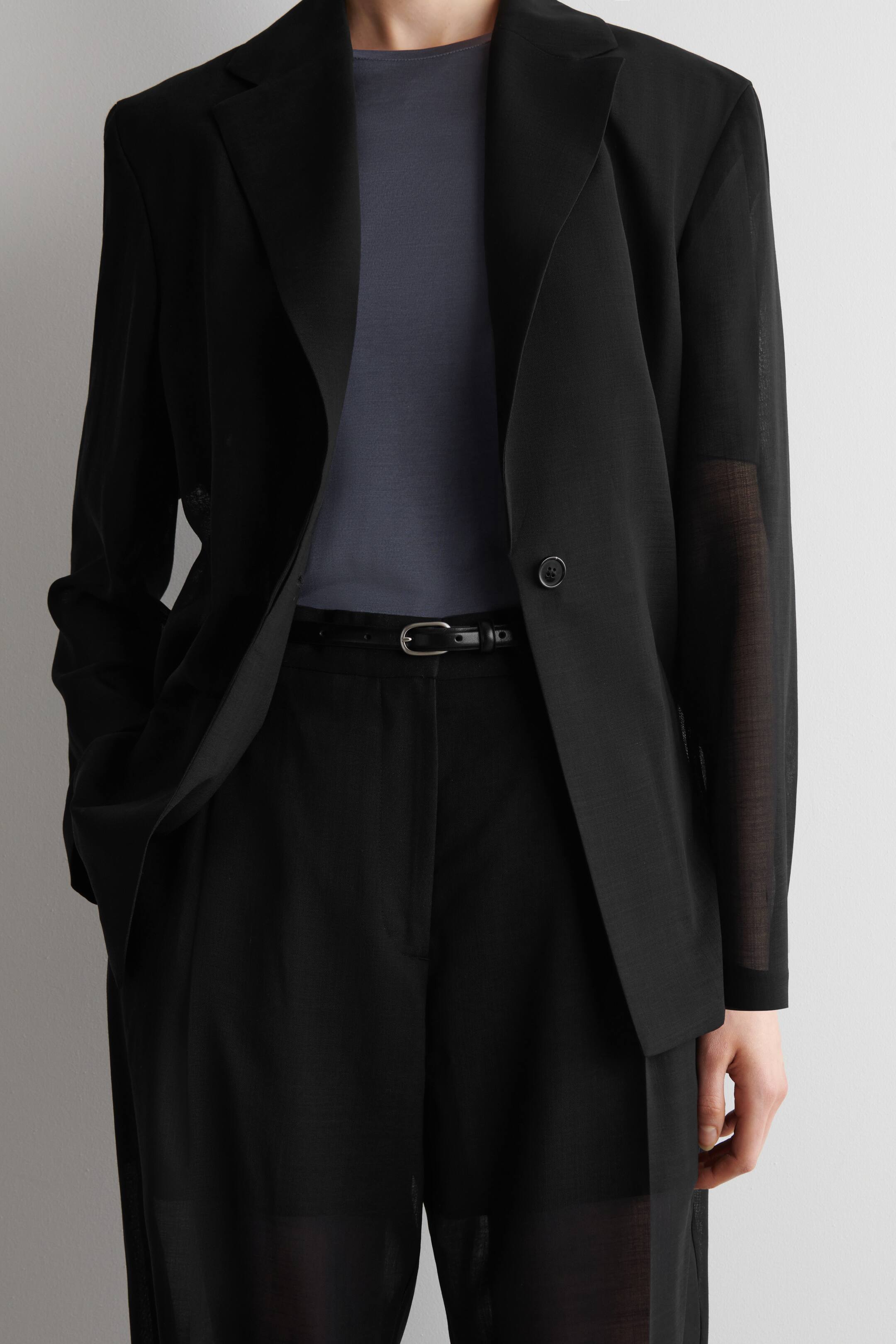 Agrandir l'image: Un blazer noir avec des revers en pointe et des manches longues transparentes est porté ouvert sur un haut à col rond gris foncé. Il est associé à un pantalon noir plissé, maintenu par une fine ceinture noire avec une boucle argentée, le tout présenté de face.