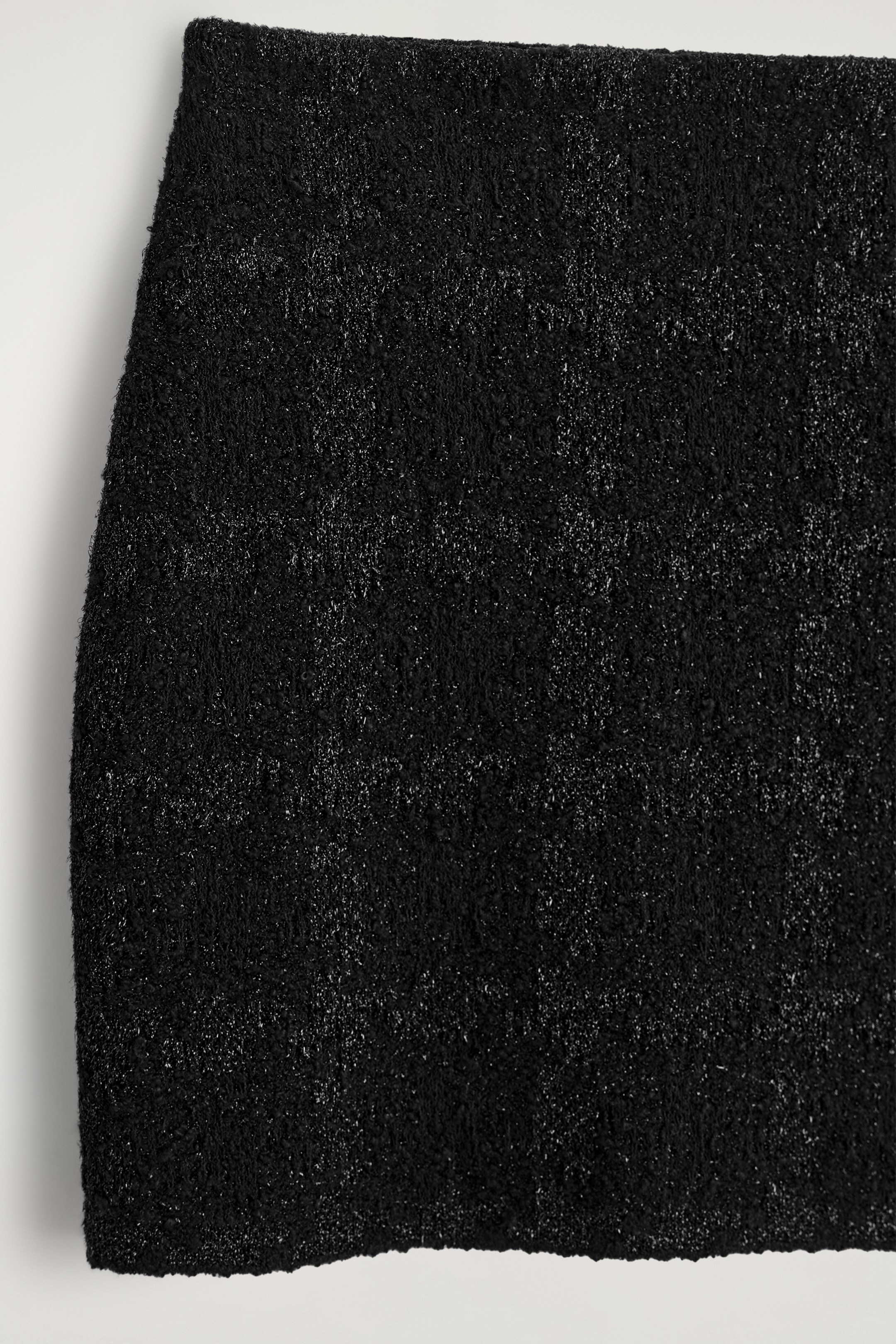 Grotere afbeelding bekijken: Een zwarte minirok wordt gepresenteerd, met een bouclé breisel dat metallic draden bevat die een subtiel ruitpatroon vormen.