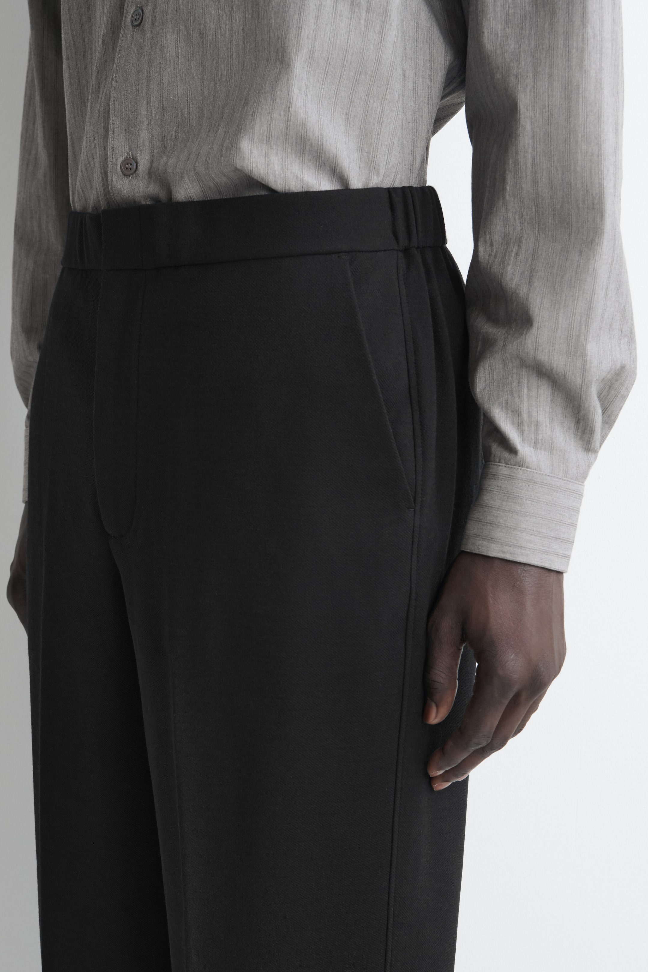 Black trousers with a structured design, a flat-front waistband with elasticated sides, and a side slip pocket are shown from a close-up, three-quarter angle. A gray, long-sleeve button-up shirt is tucked into the waistband, highlighting the trousers’ clean fit and structure. Black trousers with a structured design, a flat-front waistband with elasticated sides, and a side slip pocket are shown from a close-up, three-quarter angle. A gray, long-sleeve button-up shirt is tucked into the waistband, highlighting the trousers’ clean fit and structure.