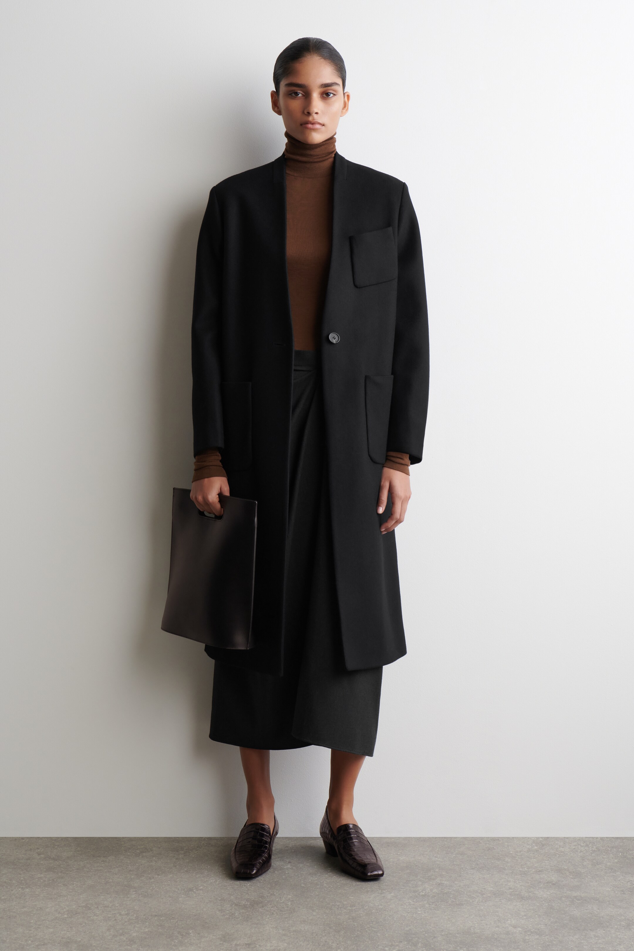 Ver imagen más grande: Un abrigo negro hasta la rodilla se presenta desde el frente, con un escote en V sin cuello y un cierre de un solo botón. Muestra un bolsillo de parche en el pecho izquierdo y dos bolsillos de parche más grandes en la parte inferior delantera. Un jersey de cuello alto marrón está en capas debajo, y una falda negra se extiende por debajo del dobladillo del abrigo.