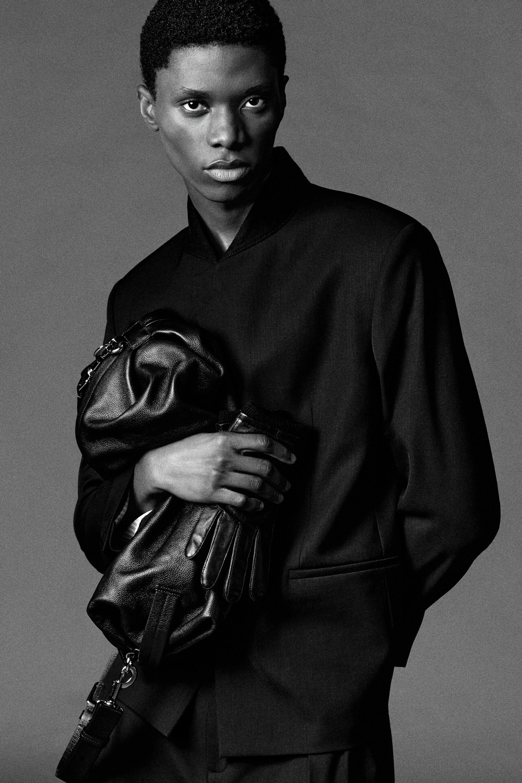 The black grained leather duffle bag is being held in a folded, slouched position by a model facing forward, who is also wearing black leather gloves and a structured black jacket. The bag features visible silver-toned metal hardware, a zip closure, and a detachable shoulder strap hanging down. The black grained leather duffle bag is being held in a folded, slouched position by a model facing forward, who is also wearing black leather gloves and a structured black jacket. The bag features visible silver-toned metal hardware, a zip closure, and a detachable shoulder strap hanging down.