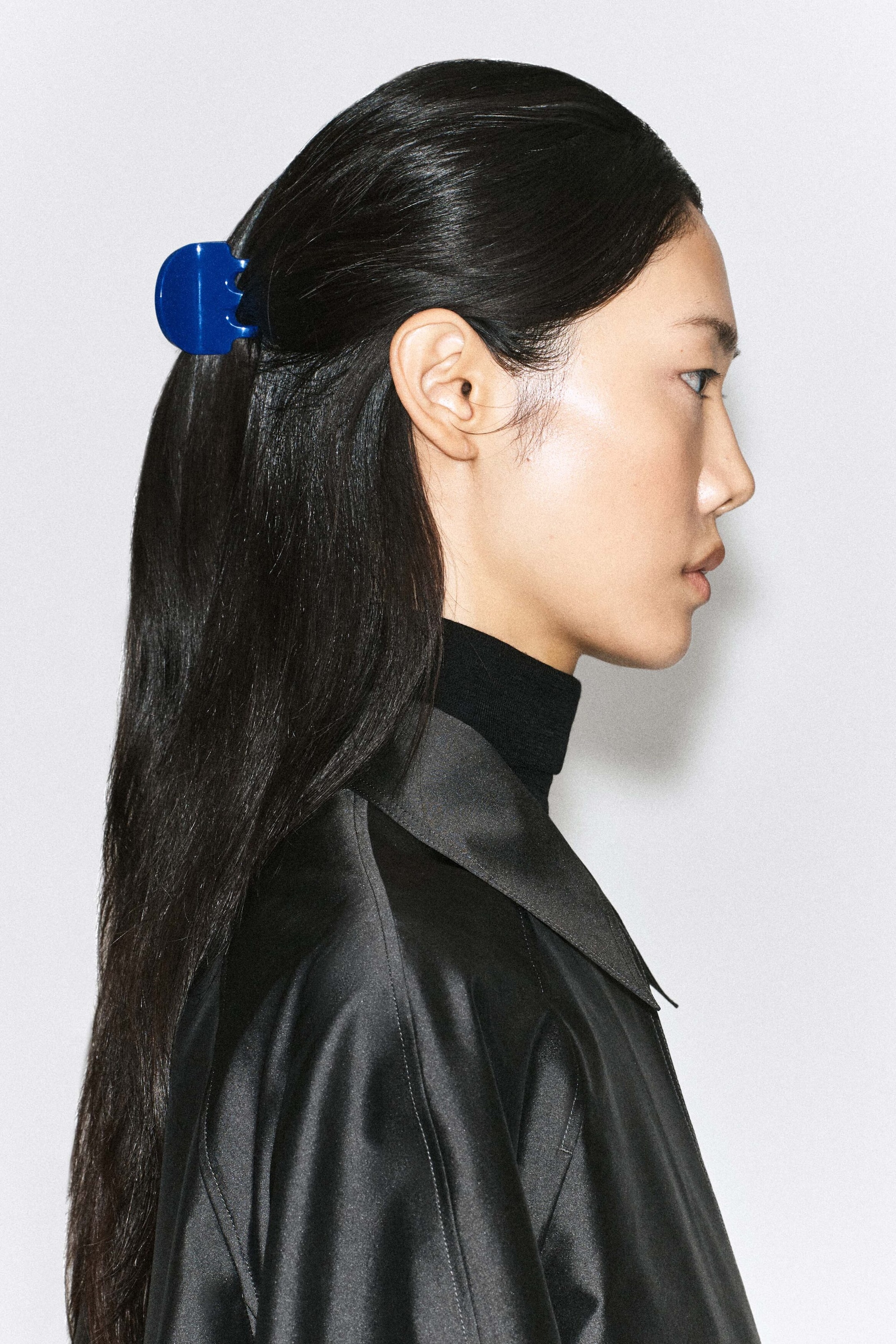 A cobalt blue hair claw clip is being worn by the model, holding a section of her long dark hair back in a half-up style. The glossy, squared clip stands out against her hair and coordinates with her black high-neck top.