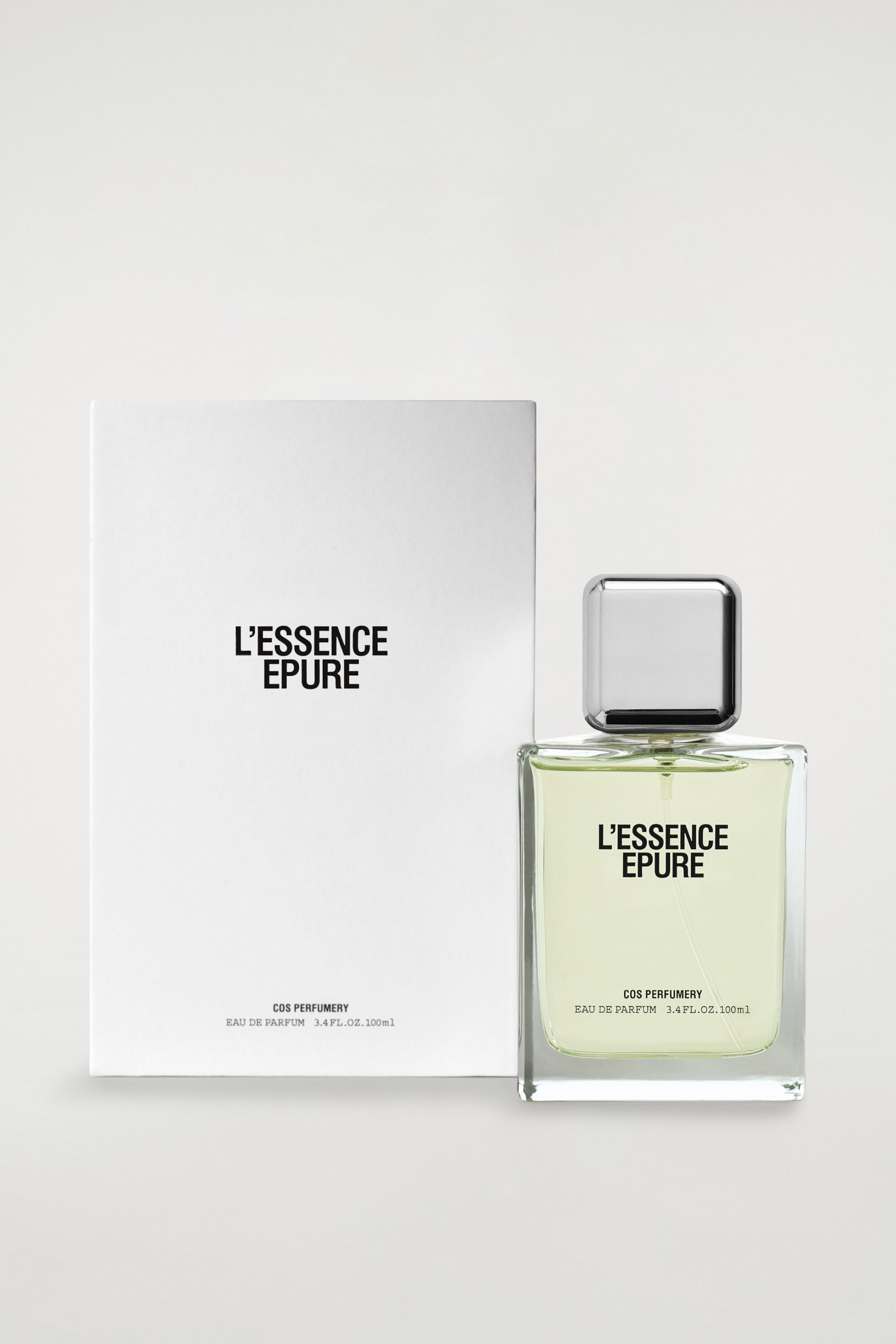 The COS Perfumery L?Essence Epure Eau De Parfum is standing facing front, featuring a clear rectangular glass bottle filled with a light green liquid and topped with a square, silver-toned cap. Standing beside the bottle is its white rectangular paper packaging box, with both items displaying black text that reads "L?ESSENCE EPURE" and "COS PERFUMERY EAU DE PARFUM 3.4 FL.OZ. 100ml." The COS Perfumery L?Essence Epure Eau De Parfum is standing facing front, featuring a clear rectangular glass bottle filled with a light green liquid and topped with a square, silver-toned cap. Standing beside the bottle is its white rectangular paper packaging box, with both items displaying black text that reads "L?ESSENCE EPURE" and "COS PERFUMERY EAU DE PARFUM 3.4 FL.OZ. 100ml."
