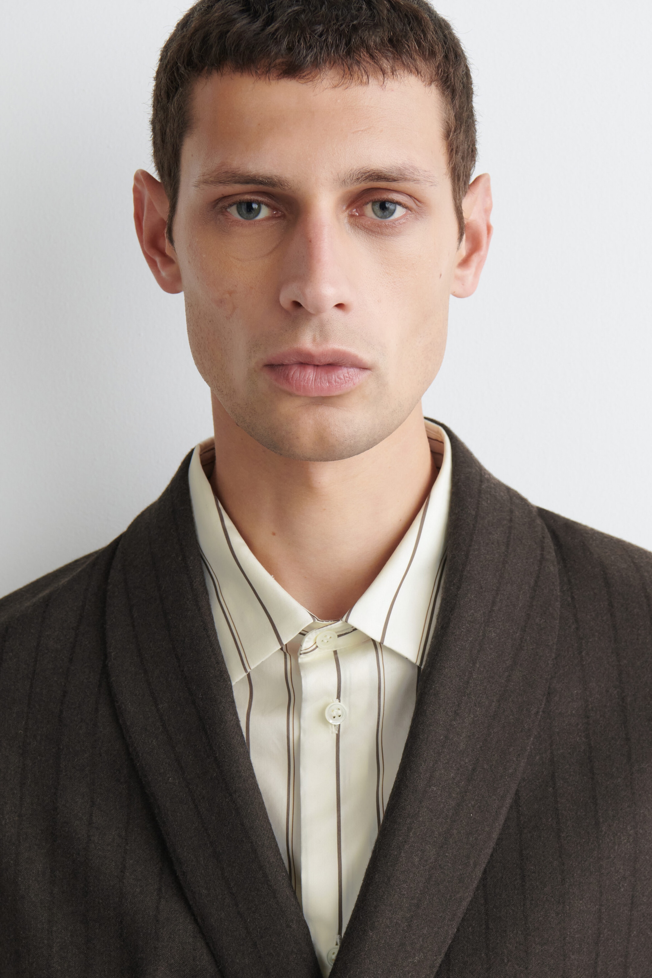 A dark brown pinstriped wool blazer is shown in a close-up view of the upper portion, highlighting its textured twill fabric and shawl collar. It is layered over a light cream, vertically striped button-up shirt. A dark brown pinstriped wool blazer is shown in a close-up view of the upper portion, highlighting its textured twill fabric and shawl collar. It is layered over a light cream, vertically striped button-up shirt.
