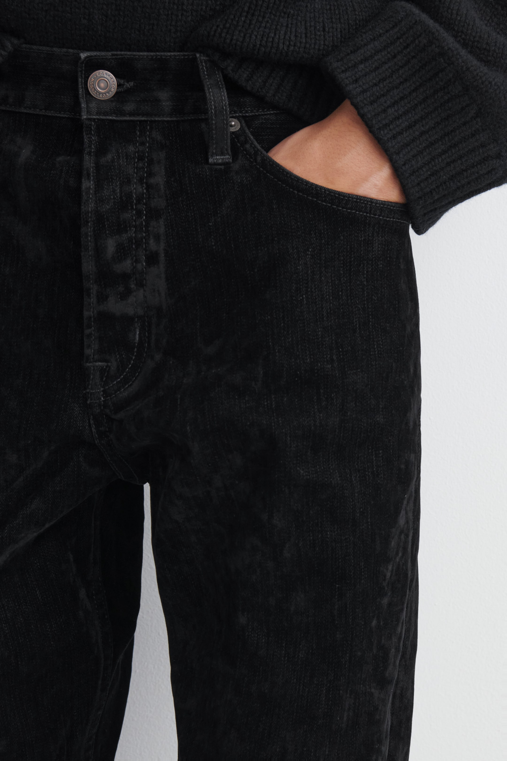 Los Signature Flocked Straight-Leg Jeans presentan una suave textura flocada negra con un acabado aterciopelado en una vista frontal de cerca. Los jeans muestran una cintura clásica con trabillas para cinturón, un botón metálico tipo shank y un bolsillo delantero con una mano descansando en su interior.