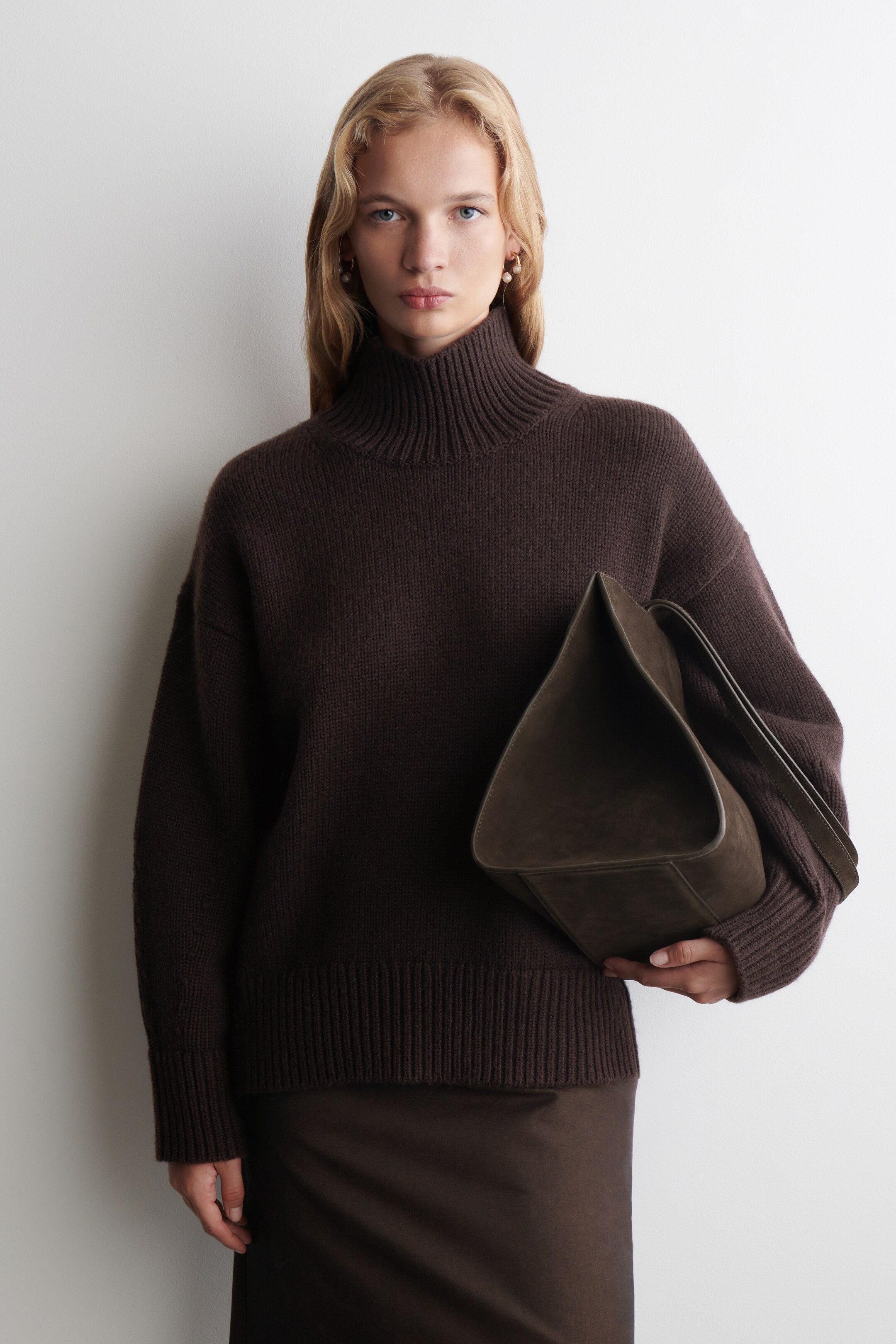 View larger image: A dark brown, knitted turtleneck sweater, featuring a prominent ribbed collar and ribbed hem, is being worn with a relaxed fit and is angled slightly to the right. A dark olive-brown, softly structured bag, appearing to be suede, is being held against the sweater in the wearer's right hand.