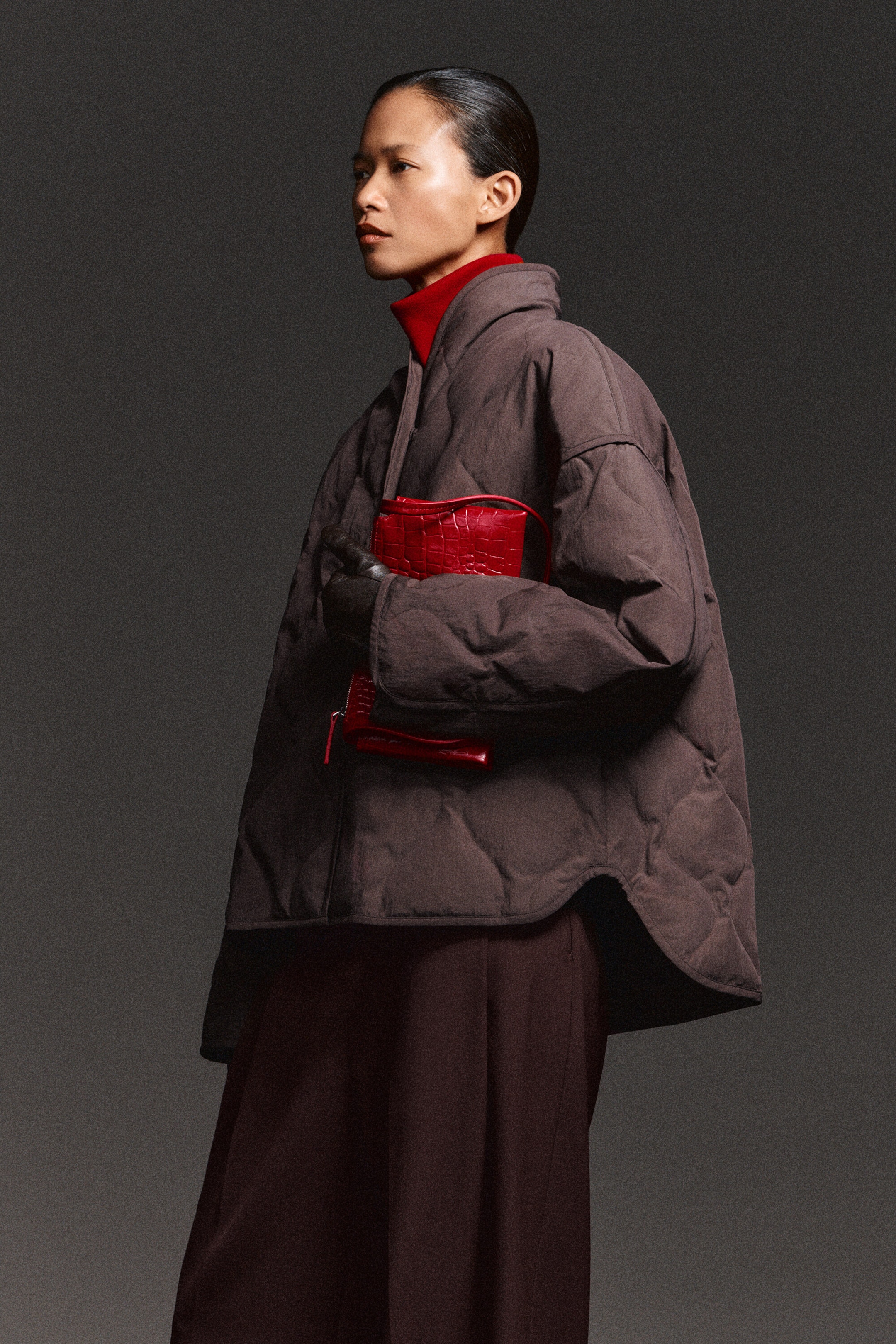 A dark brown shawl-collar quilted jacket is shown from a three-quarter angle, highlighting its oversized silhouette and diamond-quilted design. The jacket features a high shawl collar layered over a red turtleneck, wide long sleeves, and a dramatic curved high-low hemline.