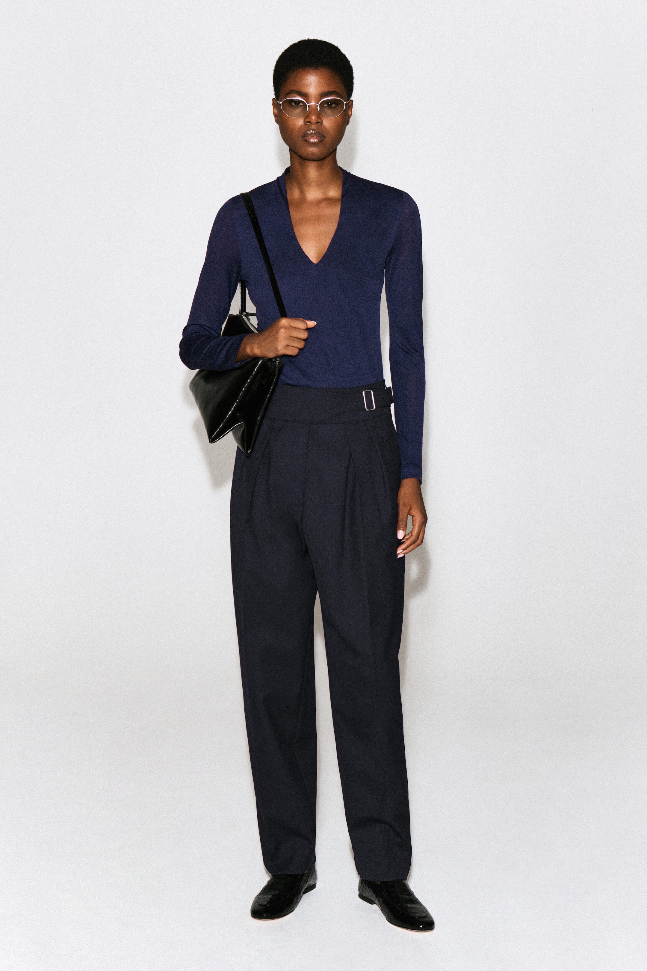 The model is wearing a dark blue, long-sleeved V-neck top tucked into high-waisted pleated trousers. The slim-fitting top is styled with a black leather shoulder bag. The model is wearing a dark blue, long-sleeved V-neck top tucked into high-waisted pleated trousers. The slim-fitting top is styled with a black leather shoulder bag.