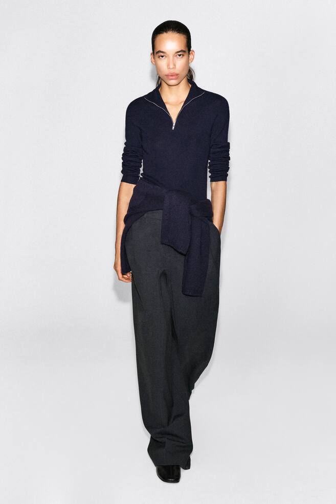 A navy blue, fine-knit half-zip sweater is shown from the front, featuring a silver-toned zip at the collar and long sleeves pushed up to the forearms. The sweater is styled with a matching navy knit tied around the waist and paired with dark grey wide-leg trousers. - 1