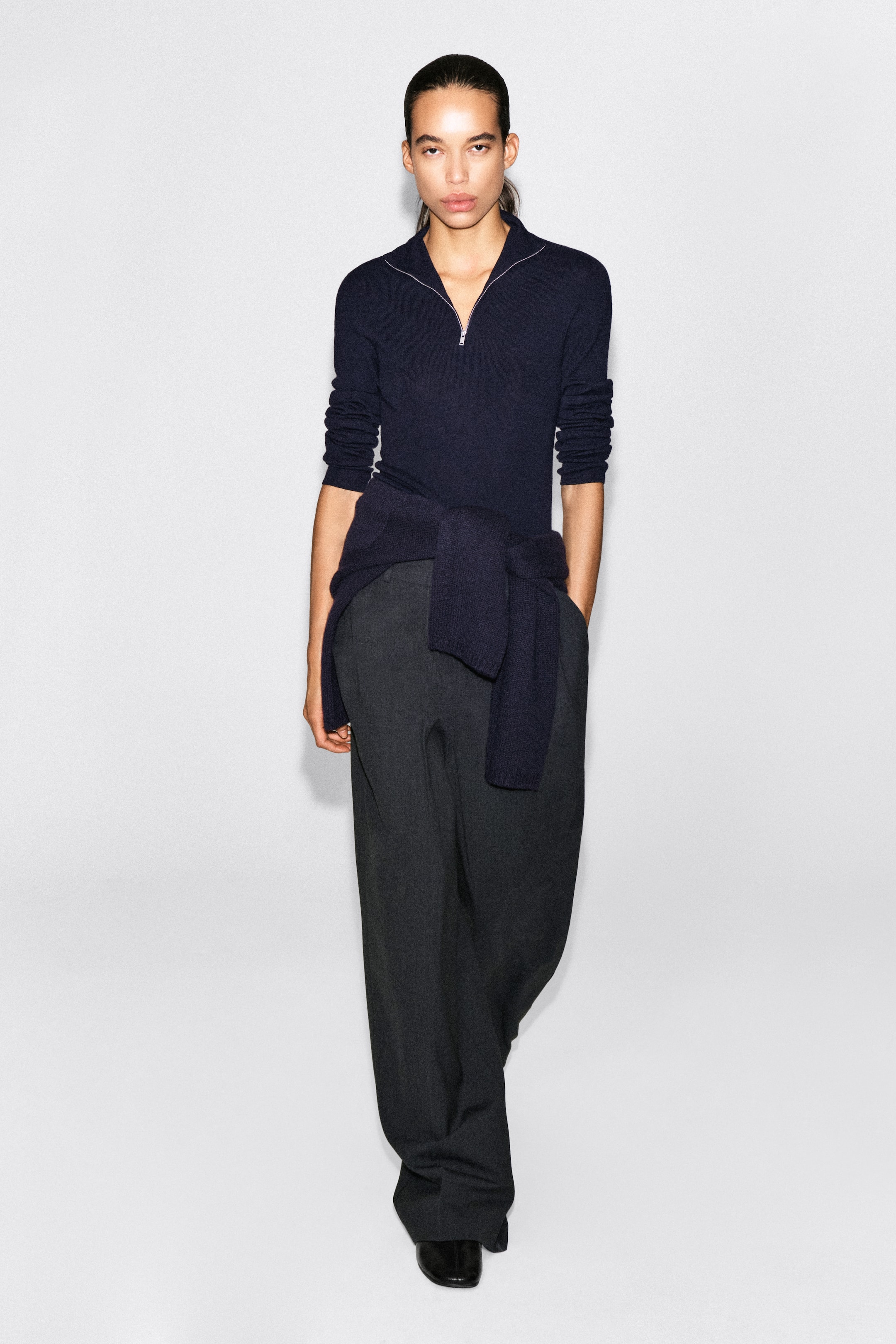 Visualizza l'immagine ingrandita: Un maglione navy blu a mezza zip in maglia fine viene presentato frontalmente, con una cerniera color argento sul colletto e maniche lunghe arrotolate fino agli avambracci. Il maglione è abbinato a una maglia navy coordinata annodata in vita e a pantaloni a gamba larga grigio scuro.