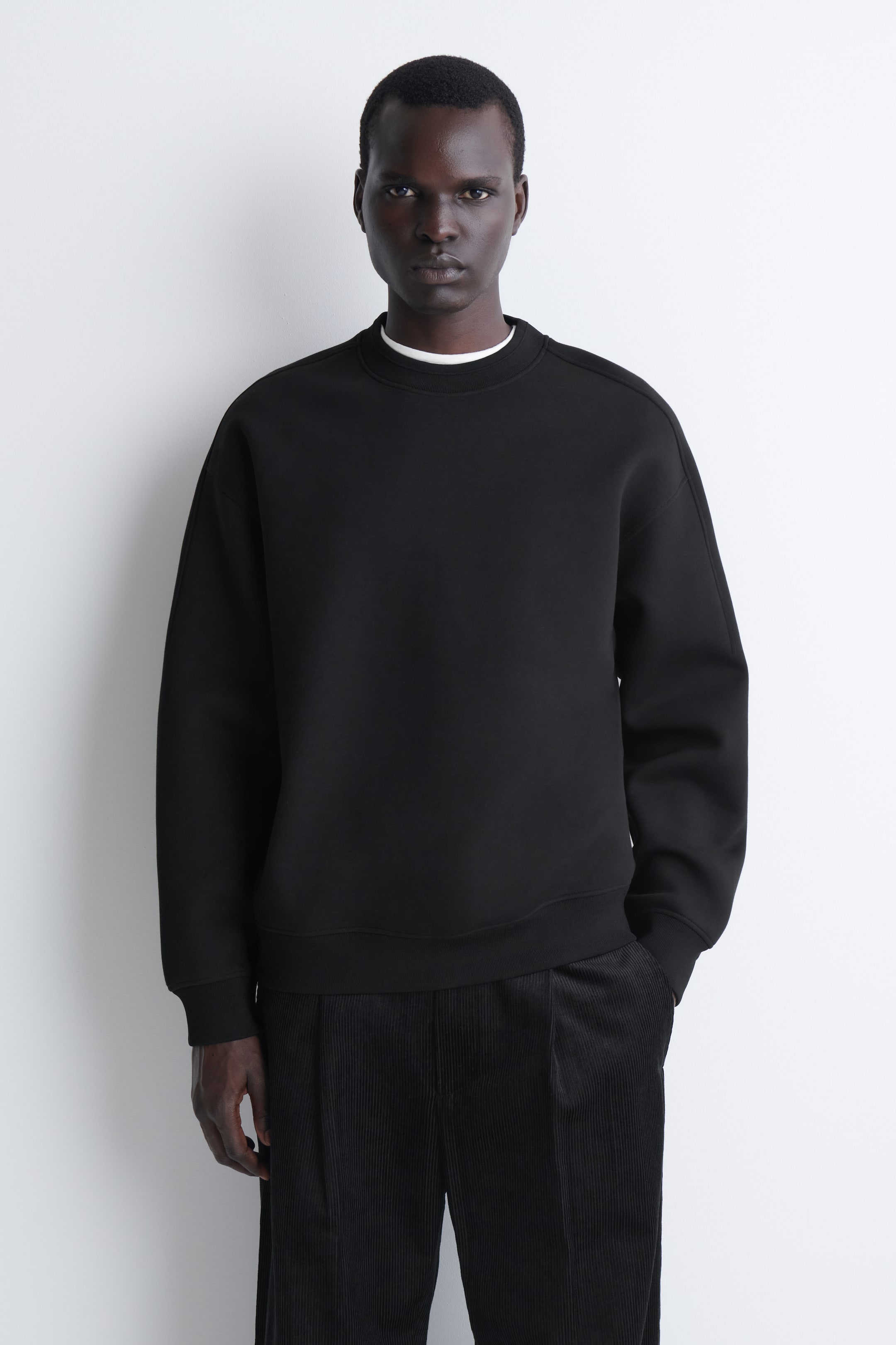 Nagyobb kép megtekintése: A fekete Relaxed Scuba Sweatshirt elölről látható, kiemelve sima, strukturált anyagát, ejtett vállait, valamint a kerek nyaknál, a mandzsettáknál és az alsó szegélynél megjelenő bordázott részleteket. A nyakkivágás alól egy fehér gallér látszik ki, amely hangsúlyozza a laza szabású darab réteges stylingját.