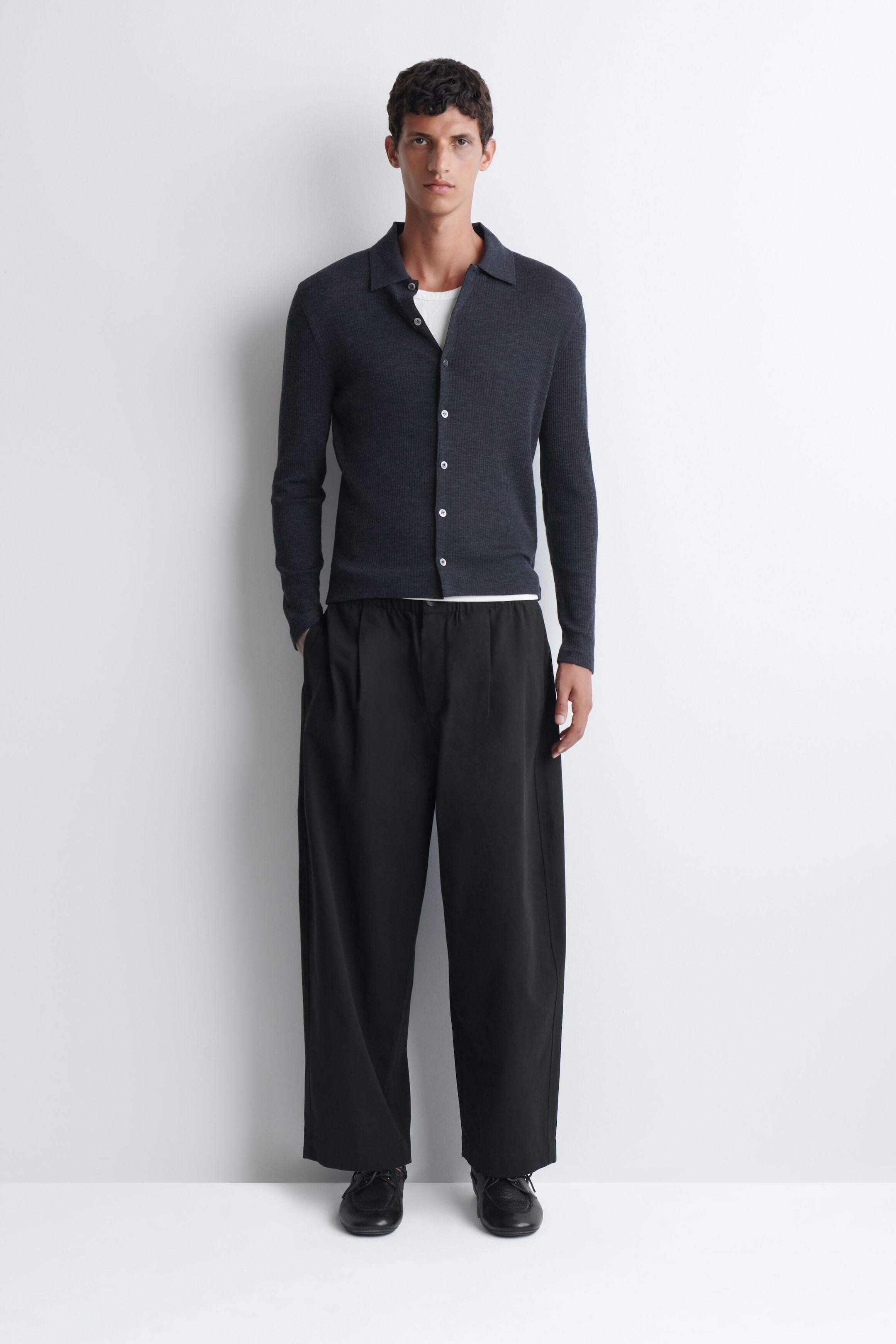 A dark grey, long-sleeved knit cardigan with a collar and white buttons is being worn by the model over a white t-shirt. The cardigan is being styled with loose-fitting, wide-leg black trousers.