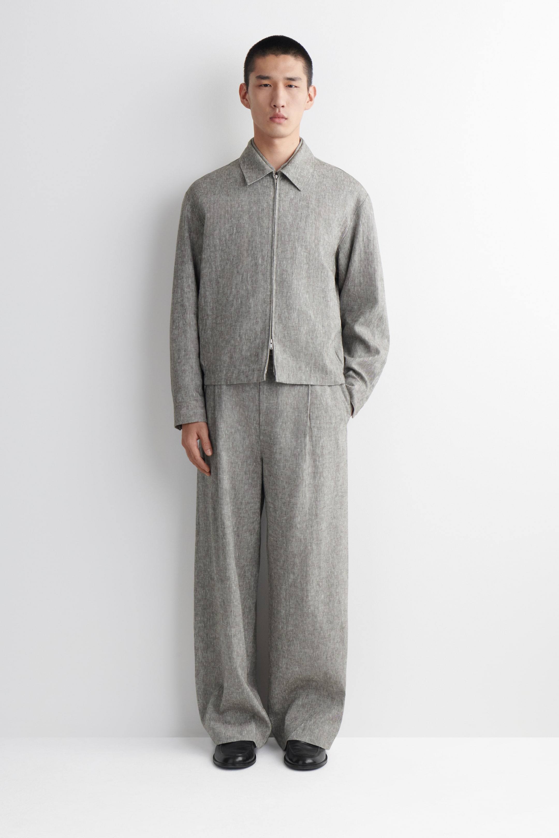 Ver imagen más grande: Se presenta un conjunto de dos piezas en gris jaspeado, que incluye una chaqueta corta con cuello, cierre frontal de cremallera completa y bolsillos laterales, combinado con pantalones de pierna ancha que tienen pliegues frontales.