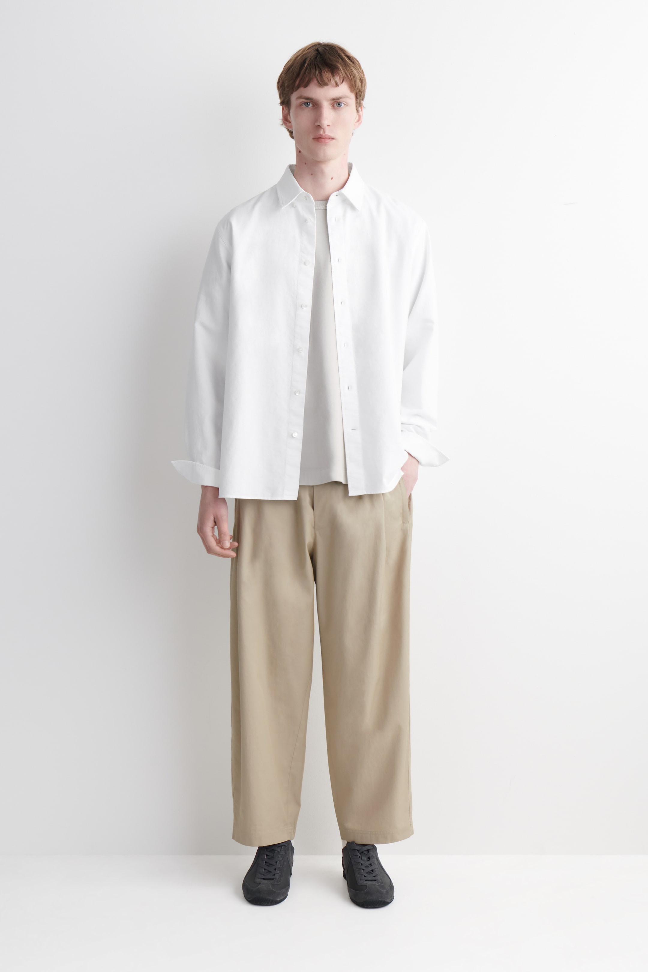 הצגת תמונה גדולה יותר: חולצה לבנה בגזרת אוברסייז עם כפתורים, בעלת צווארון פתוח ושרוולים מעט מגולגלים, נלבשת פתוחה מעל חולצת טי בצבע בז' בהיר עם צווארון עגול. ההופעה מושלמת עם מכנסיים רחבים עם קפלים בצבע חאקי ונעלי סניקרס שרוכים בצבע אפור כהה, כל זאת מוצג על אדם הפונה קדימה.