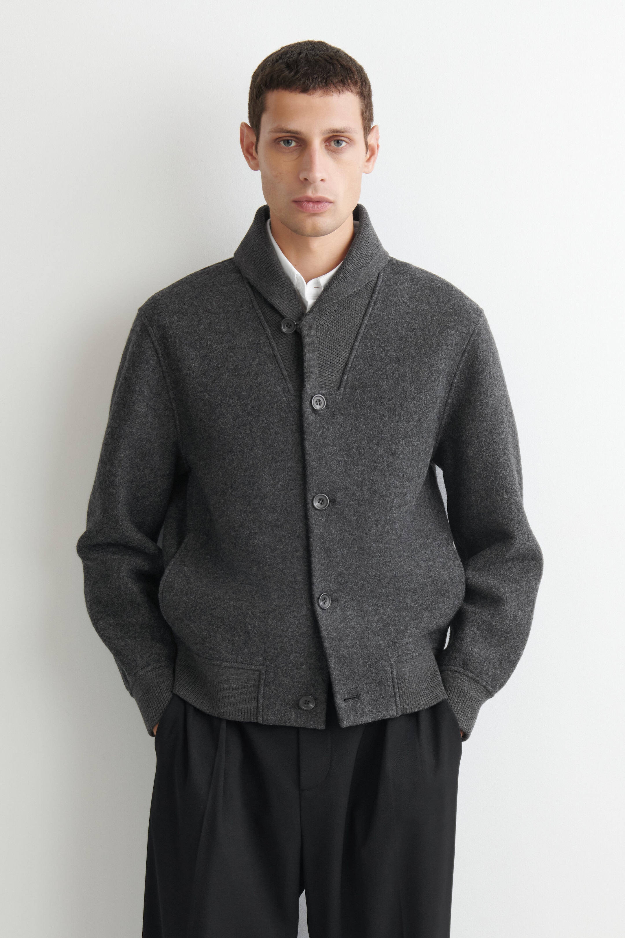 This dark gray boiled wool shawl-collar jacket is shown from the front, featuring a button-front closure, ribbed cuffs, and a ribbed hem. The jacket is layered over a white collared shirt, showcasing its relaxed yet structured fit. This dark gray boiled wool shawl-collar jacket is shown from the front, featuring a button-front closure, ribbed cuffs, and a ribbed hem. The jacket is layered over a white collared shirt, showcasing its relaxed yet structured fit.