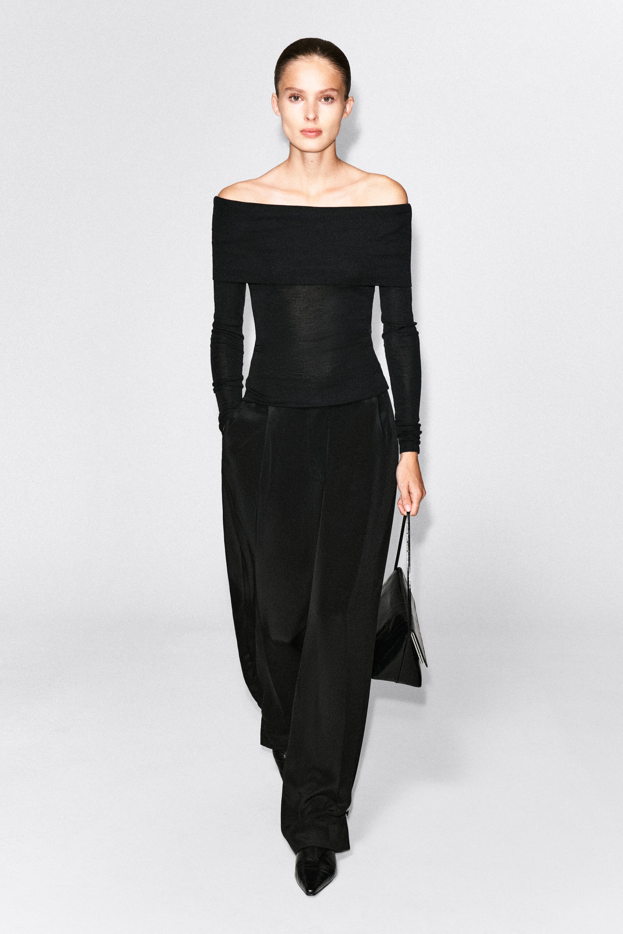 The model is wearing an off-the-shoulder black merino wool top featuring a fitted silhouette, long sleeves, and a wide fold-over neckline. The top is styled with wide-leg black trousers, and the model is holding a black handbag.