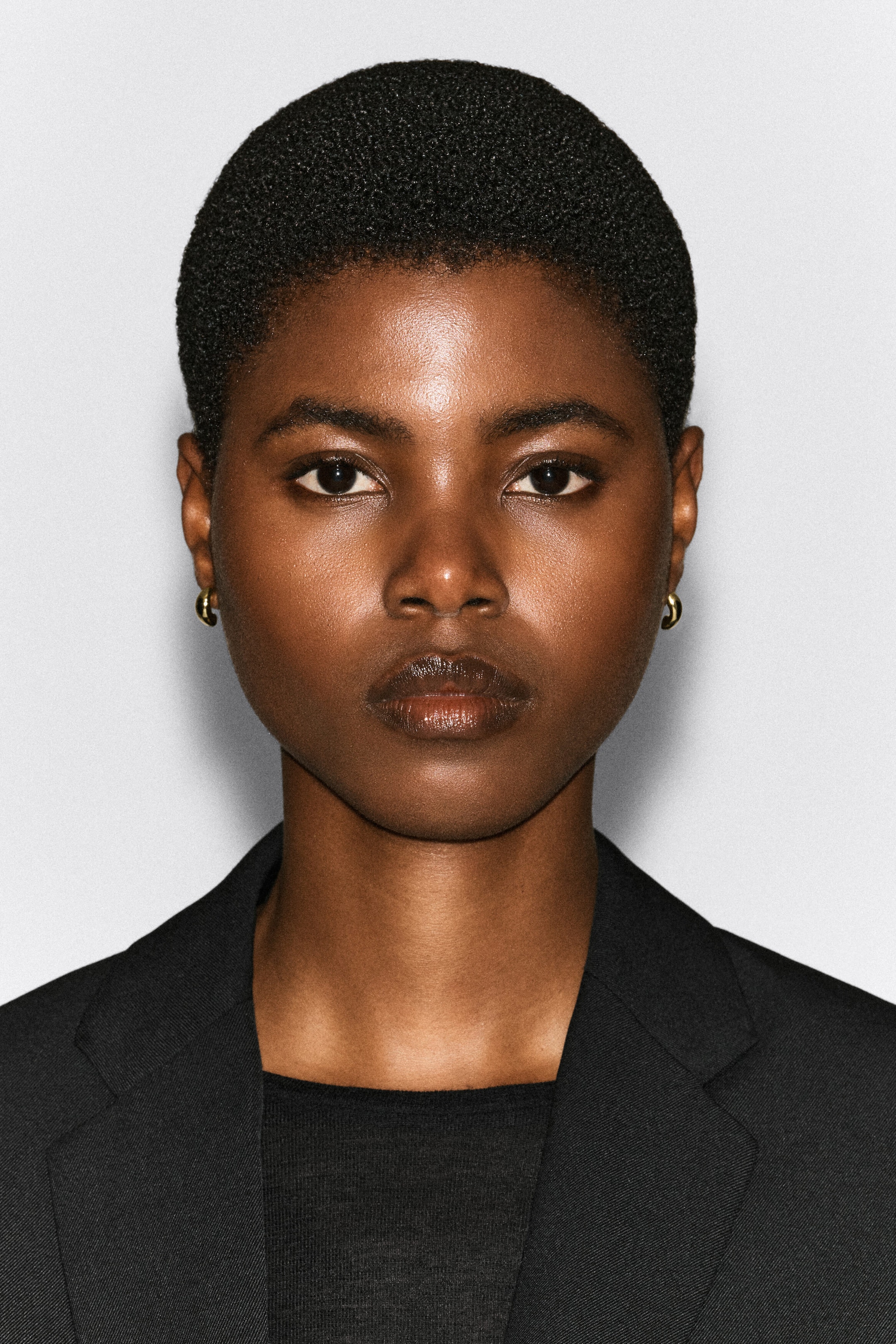 Des boucles d’oreilles demi-créoles sculpturales en métal doré poli sont portées aux lobes des oreilles du modèle. Les petites boucles d’oreilles épaisses et arrondies sont stylées avec un blazer noir et un haut assorti.