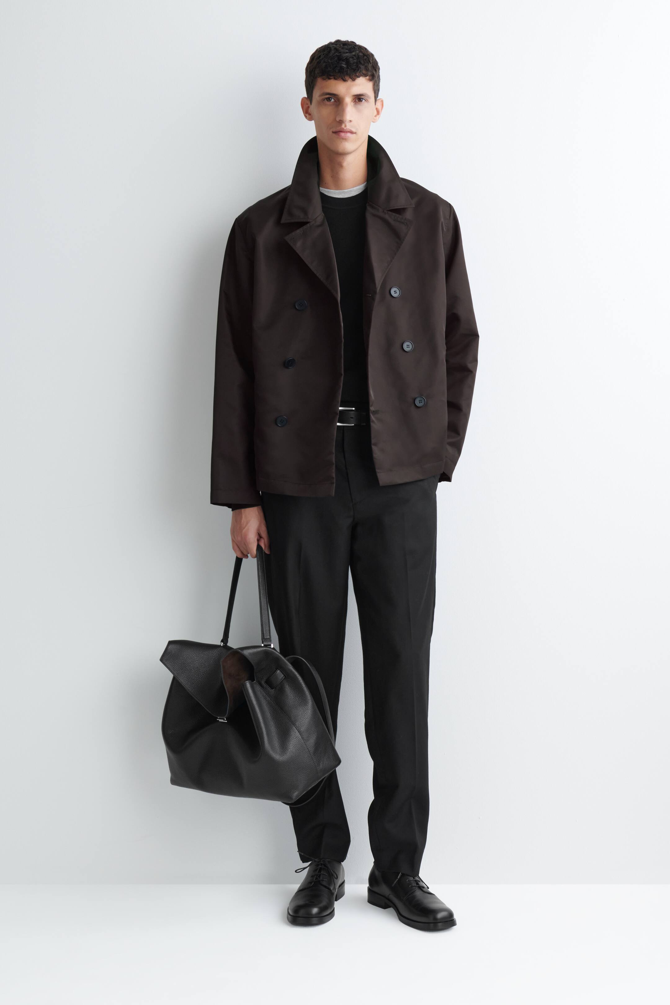 Visa större bild: En mörkbrun teknisk trenchjacka i nylon bärs uppknäppt av modellen över en svart rundhalsad tröja och svarta byxor. Jackan har en dubbelknäppt design med bred krage och stylas med en stor svart läderväska som bärs i ena handen.