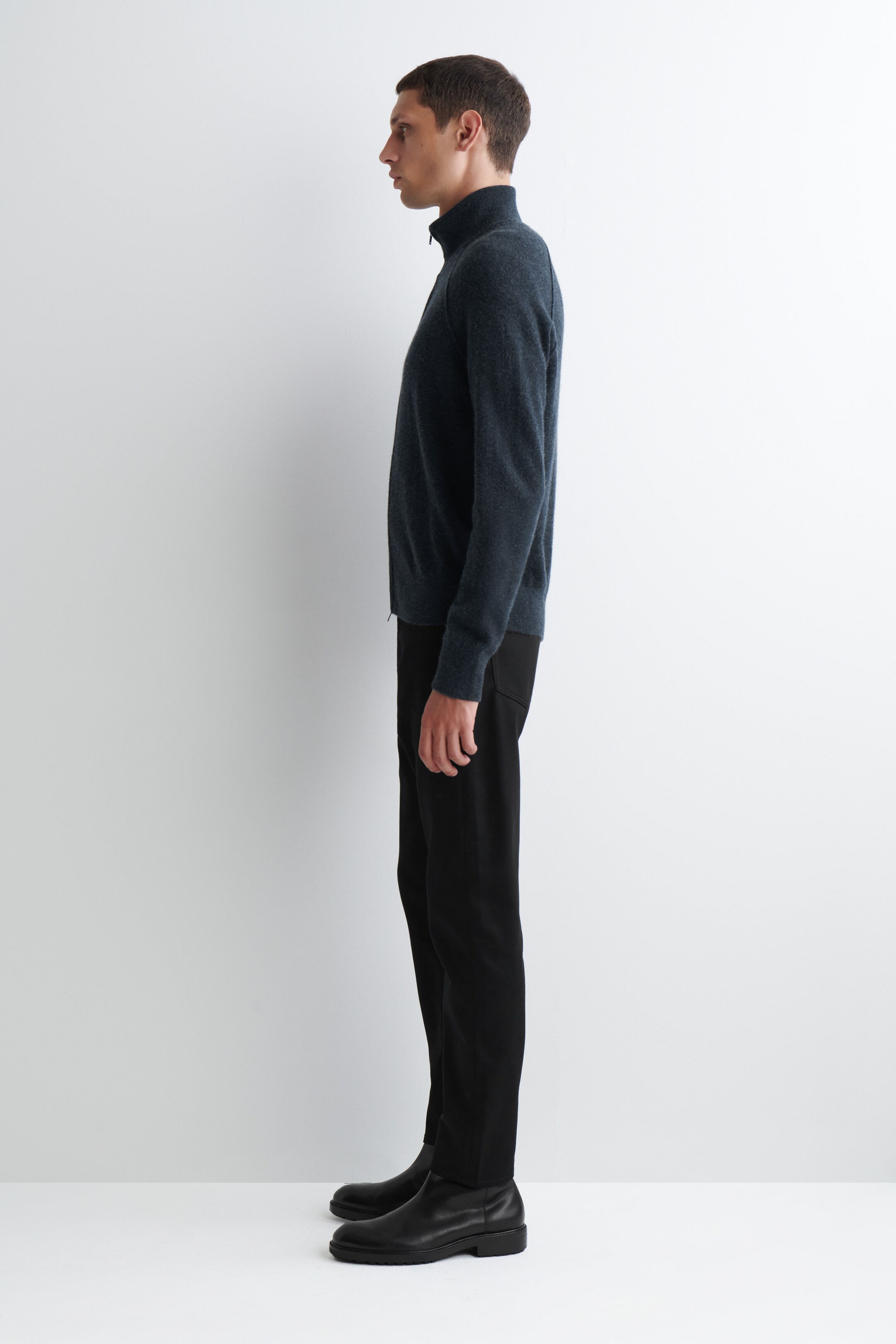 Grösseres Bild ansehen: Ein dunkelblauer Full-Zip-Pullover mit Stehkragen und gerippten Bündchen wird im Seitenprofil an einem männlichen Model präsentiert. Er wird mit schwarzen Hosen und schwarzen Lederstiefeln gestylt, um die Passform und Struktur der Kleidungsstücke zu zeigen.