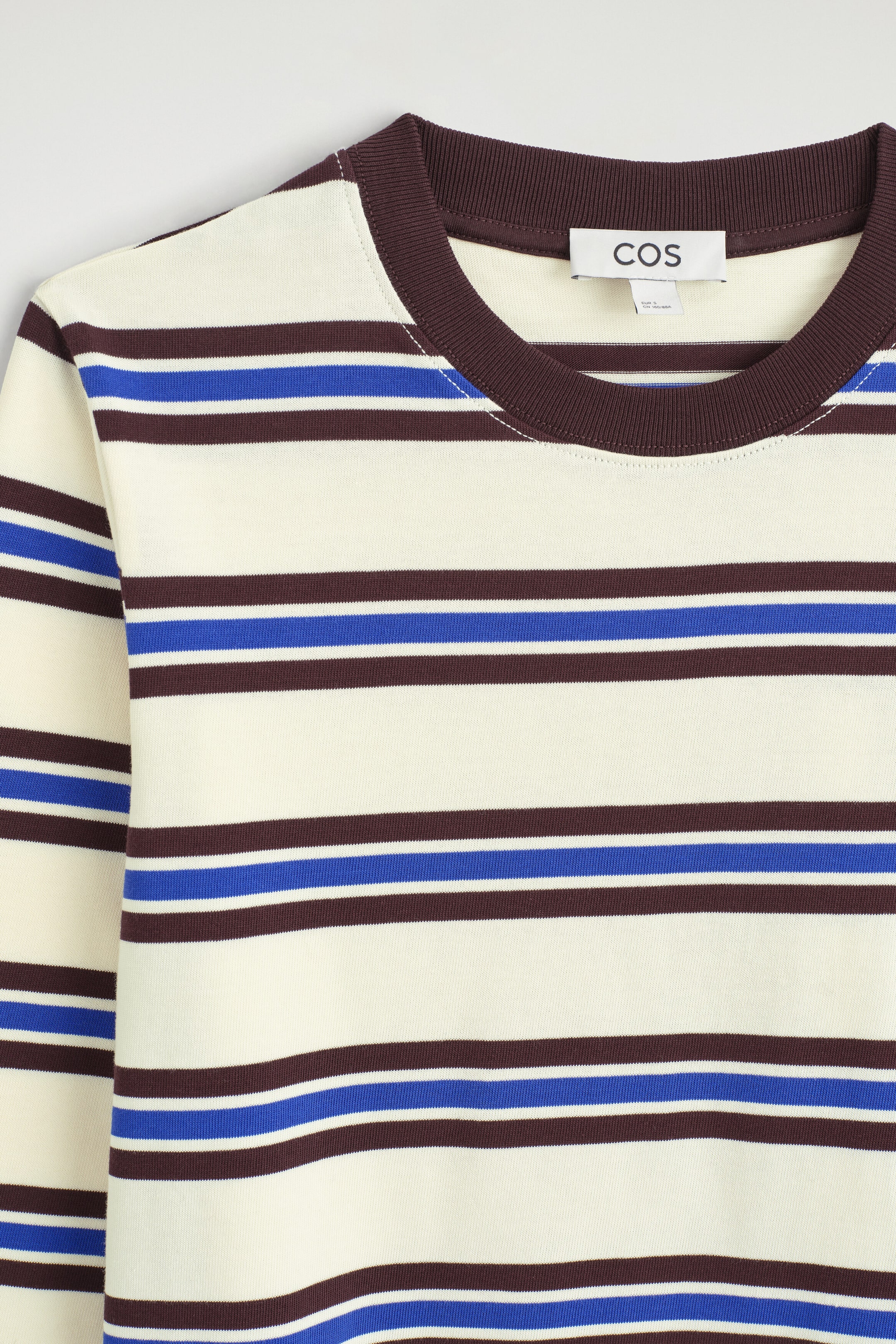 The clean-cut long-sleeved T-shirt is shown in a close-up front view, featuring horizontal stripes in cream, dark brown, and bright blue. It showcases a contrasting dark brown ribbed crew neck, with a brand tag visible on the inside collar. The clean-cut long-sleeved T-shirt is shown in a close-up front view, featuring horizontal stripes in cream, dark brown, and bright blue. It showcases a contrasting dark brown ribbed crew neck, with a brand tag visible on the inside collar.