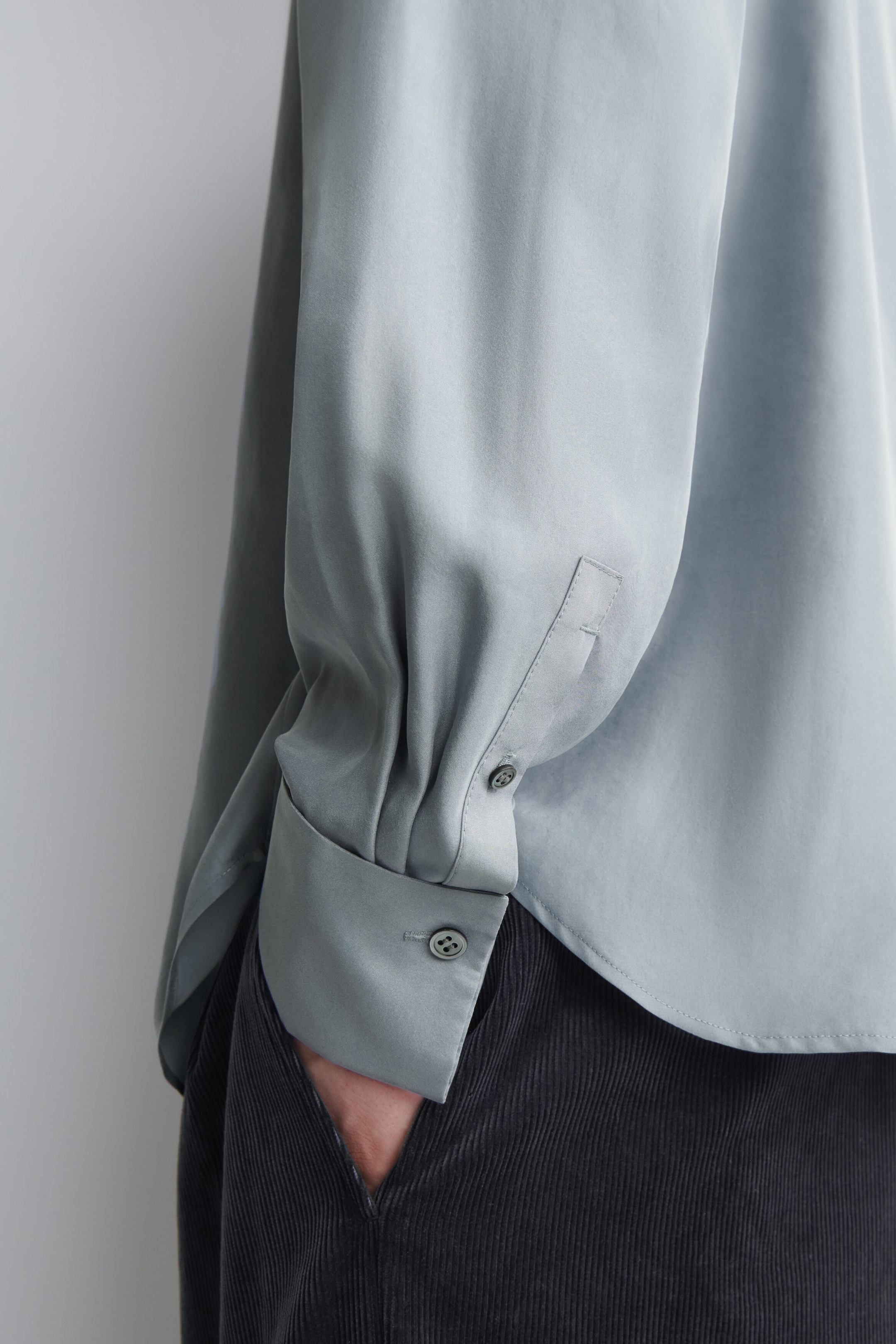 A relaxed silk shirt in a light grey-green colour is being worn by the model, featuring a pleated sleeve with a buttoned cuff and a curved hem. The shirt is styled with dark corduroy trousers, with the model’s hand partially tucked into a pocket. A relaxed silk shirt in a light grey-green colour is being worn by the model, featuring a pleated sleeve with a buttoned cuff and a curved hem. The shirt is styled with dark corduroy trousers, with the model’s hand partially tucked into a pocket.